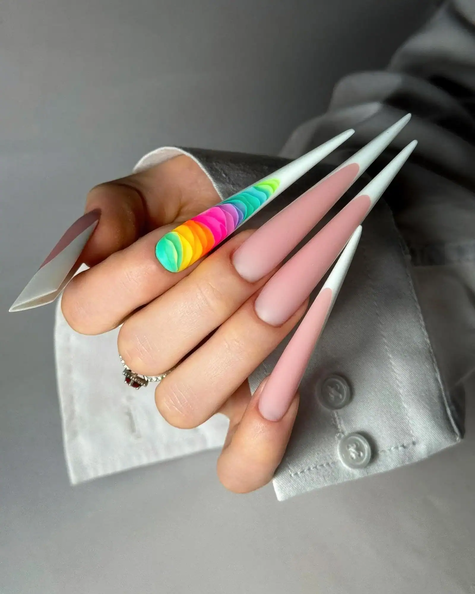 A hand holding up very long stiletto nails in soft pink with white tips and one rainbow swirl nail
