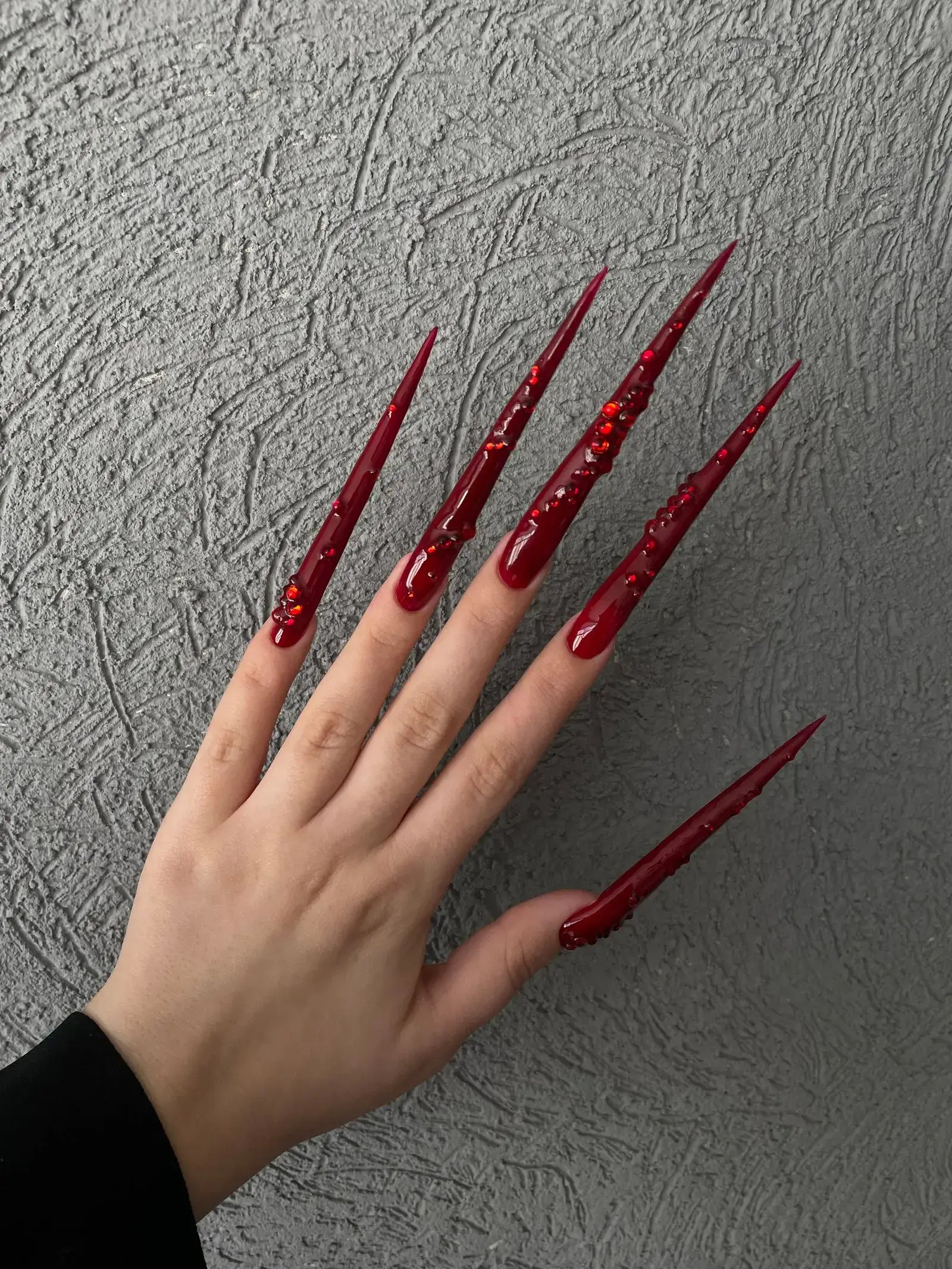 A hand with extremely long red stiletto nails with raised details on a textured grey surface