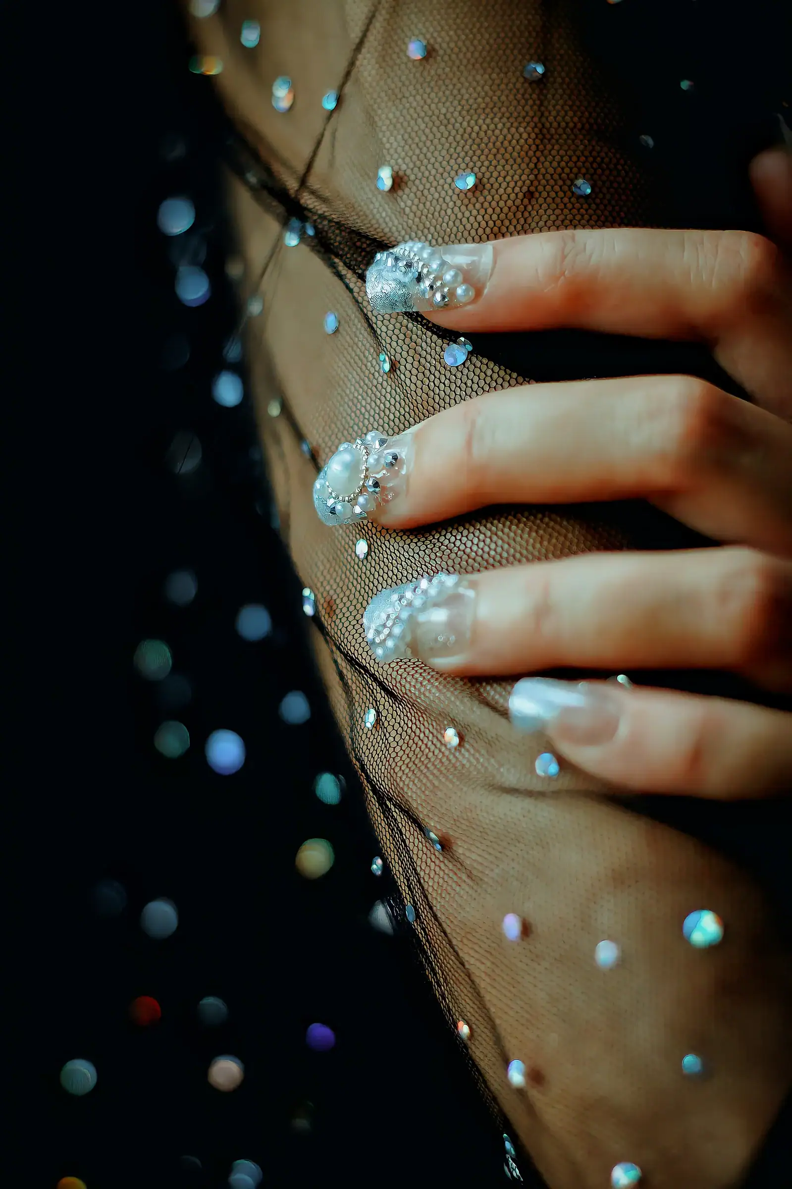 A hand with crystal-covered nails resting on black mesh scattered with crystals