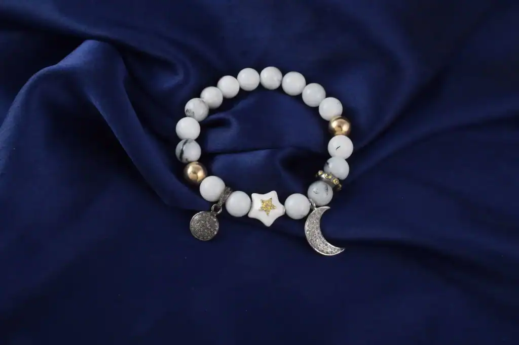 A white beaded bracelet with a star charm, a crescent moon charm and a round charm on deep blue silk.