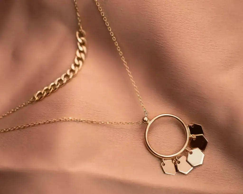 Layered gold chains, one with a round link holding small hexagonal charms, on soft pink fabric.