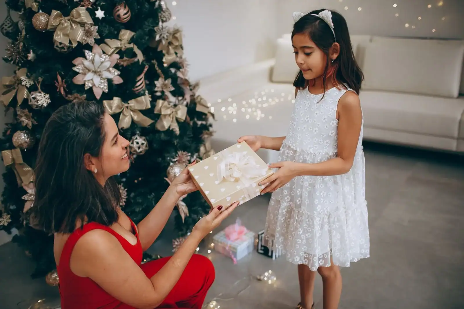 A mother in a red dress handing a wrapped gift to her daughter beside a Christmas tree