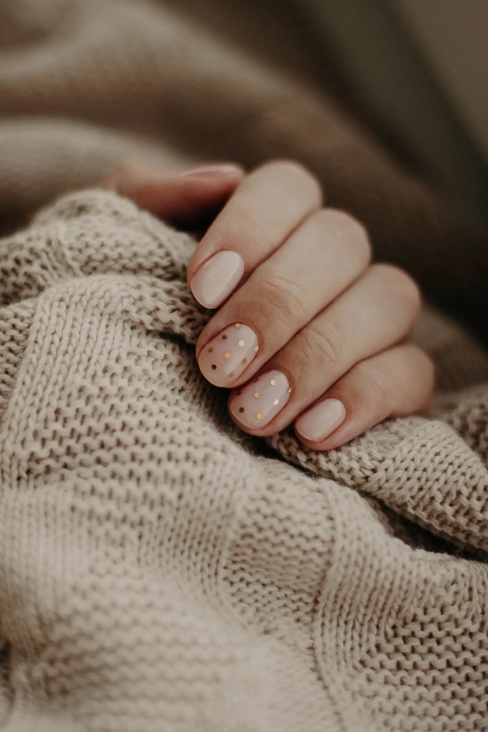Short nude nails with tiny dark dots on one nail, resting on a beige knit sweater