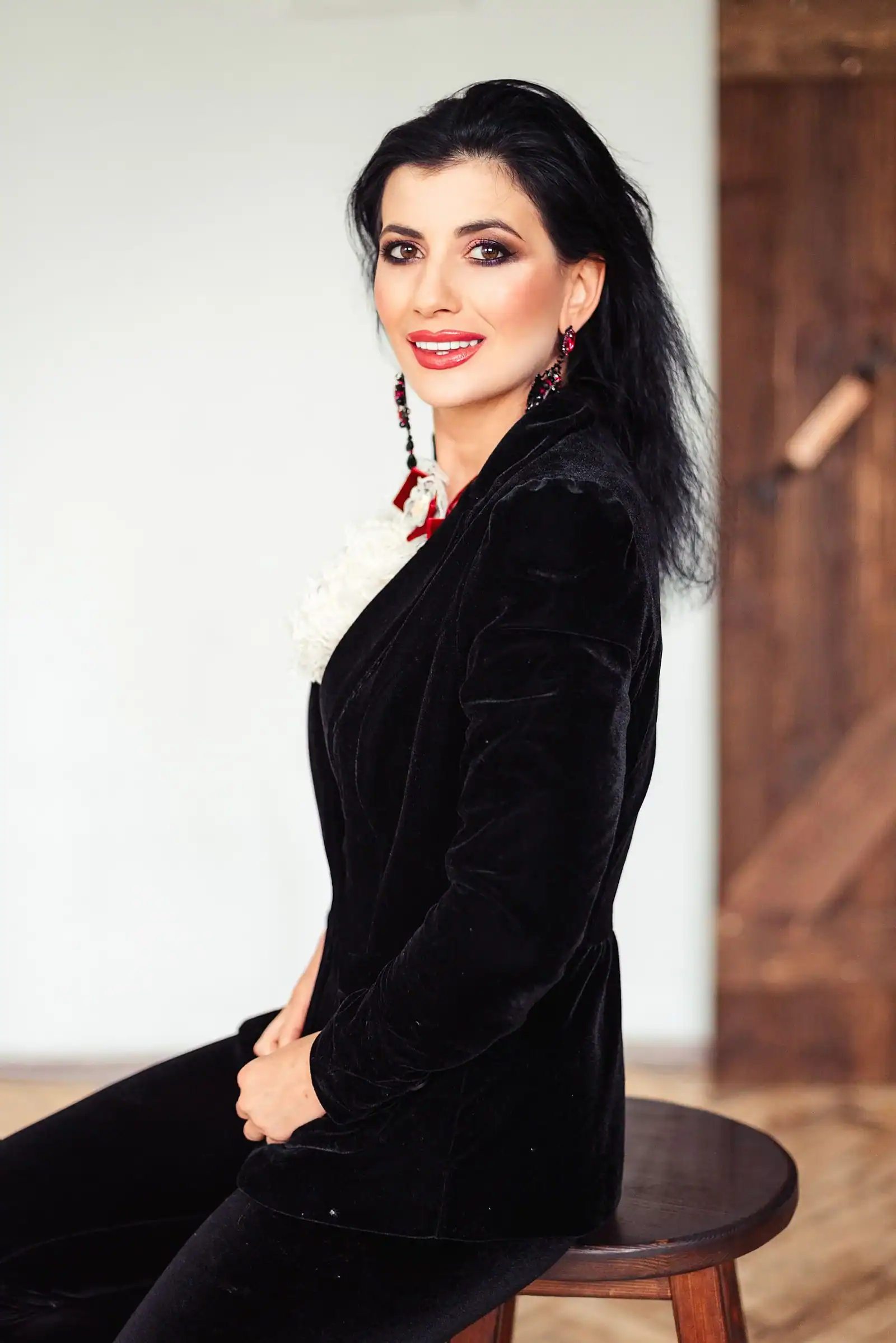 A woman in a black velvet blazer and dark trousers sits on a wooden stool