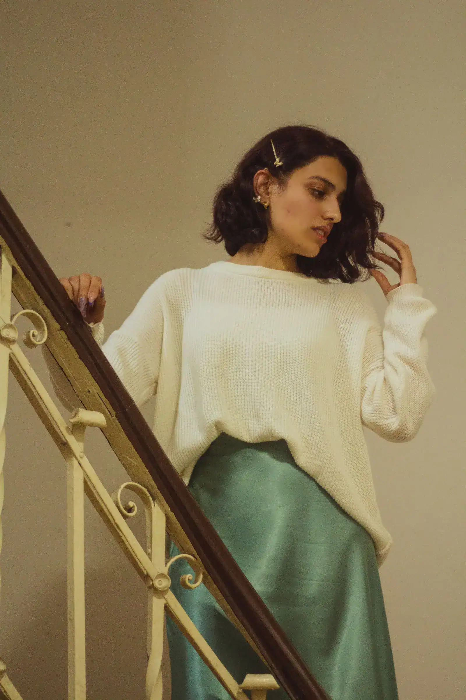 A woman in an oversized cream sweater and a green satin midi skirt on a staircase