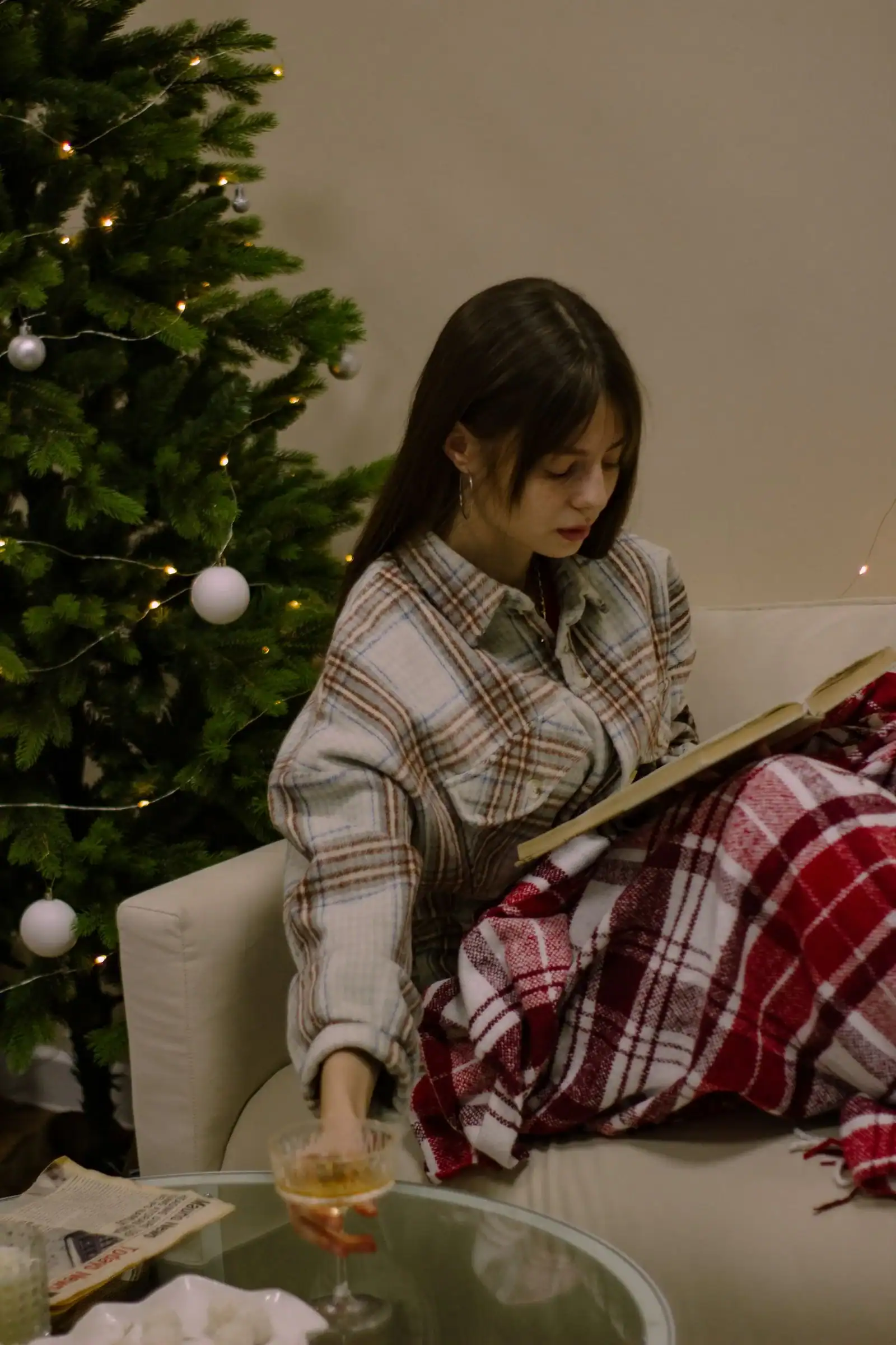 A woman in a plaid flannel shirt reads on a sofa beside a Christmas tree
