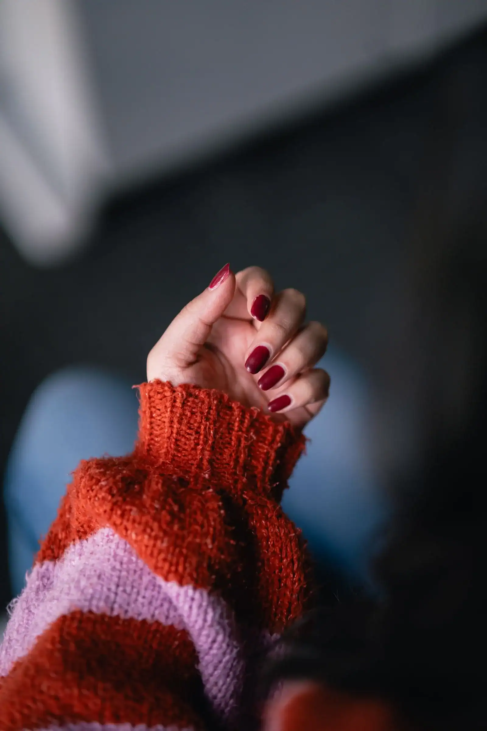 Short glossy burgundy nails peeking out of a red and pink striped sweater sleeve