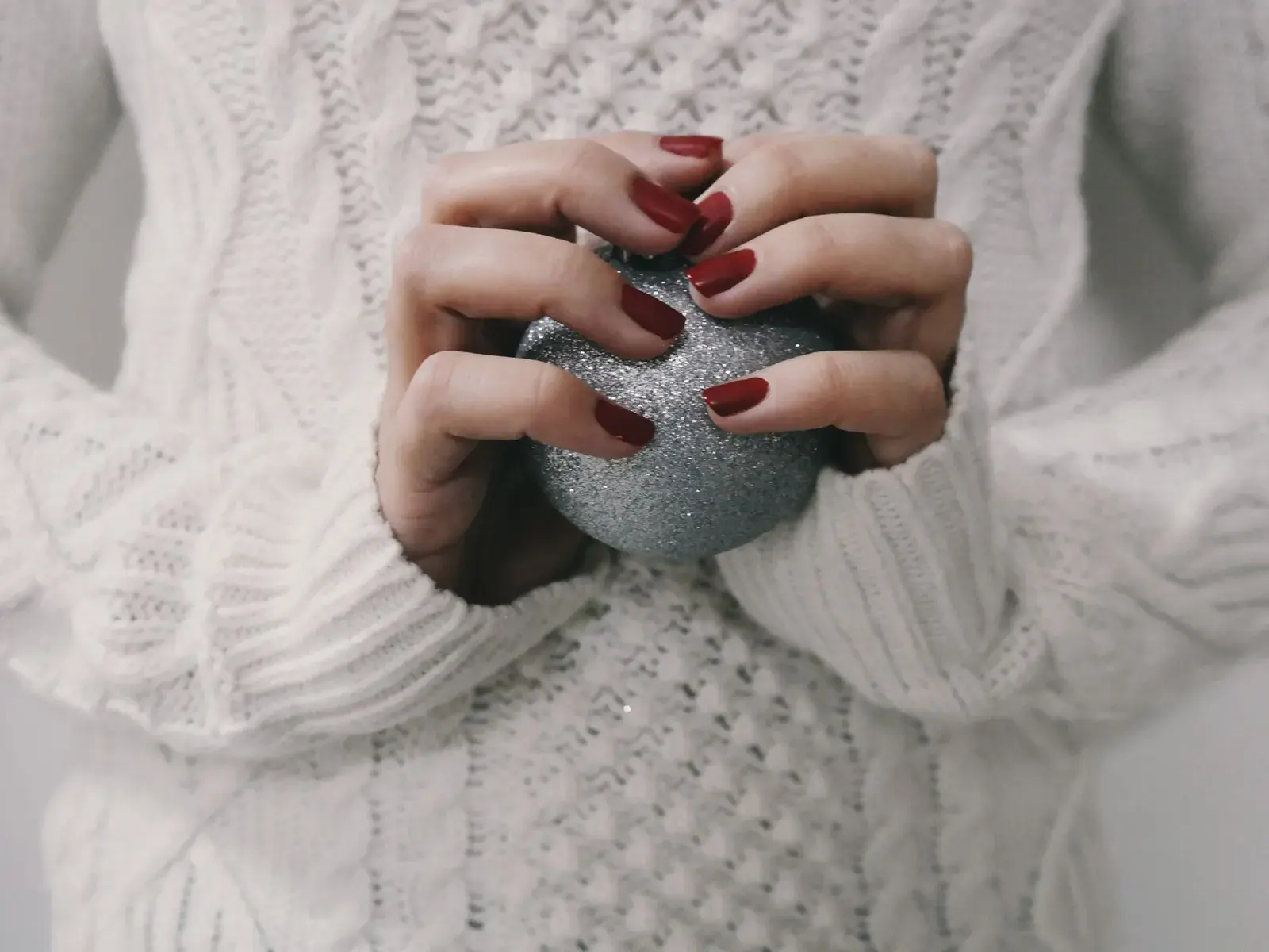 Glossy wine red nails holding a silver glitter ornament against a white cable-knit sweater