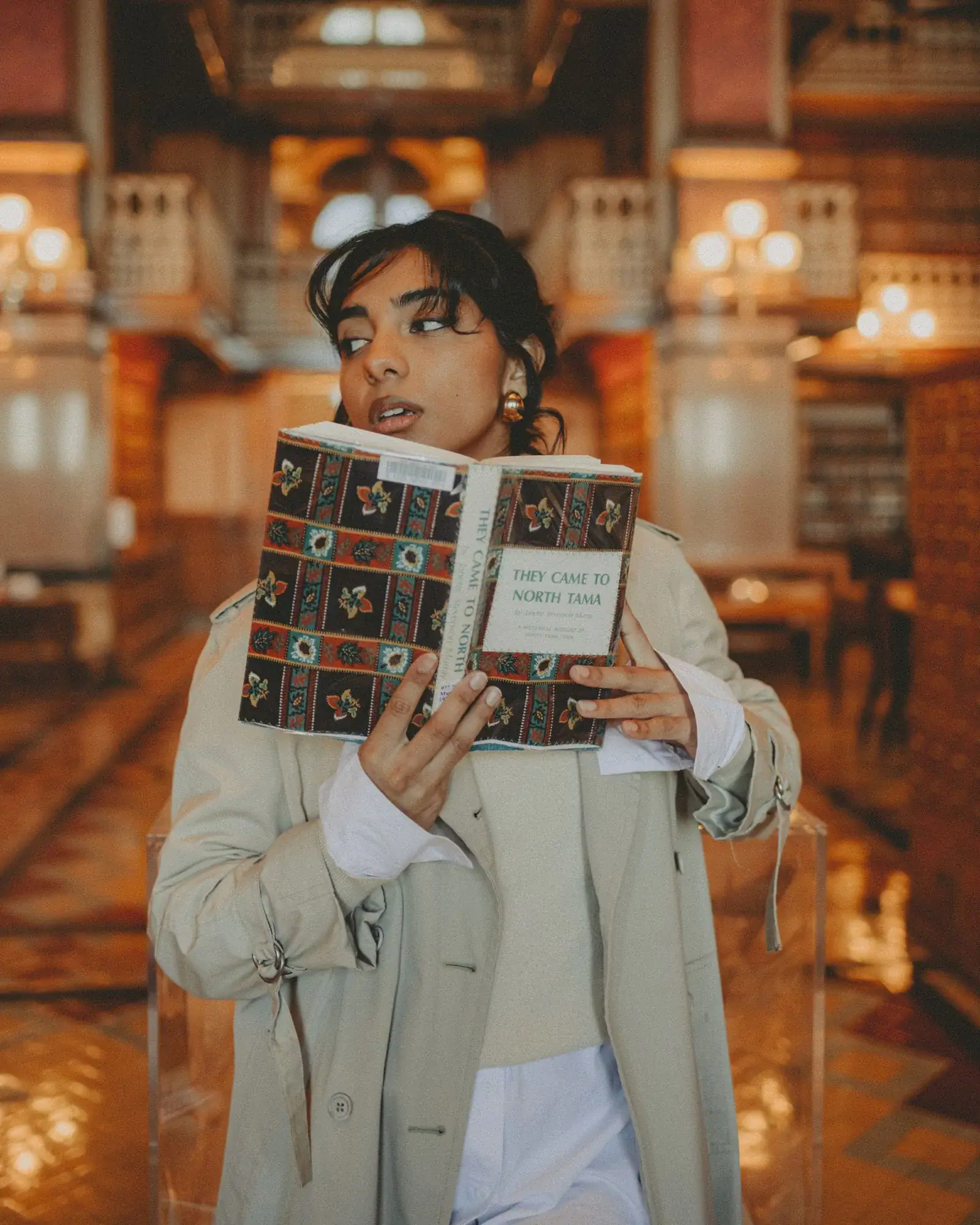 A woman in a beige coat reading an open book in an old library
