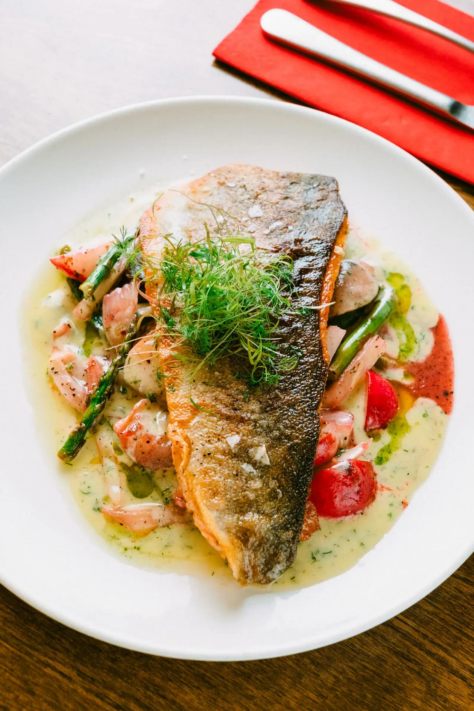 A pan-seared salmon fillet topped with dill on a white plate with asparagus and cream sauce