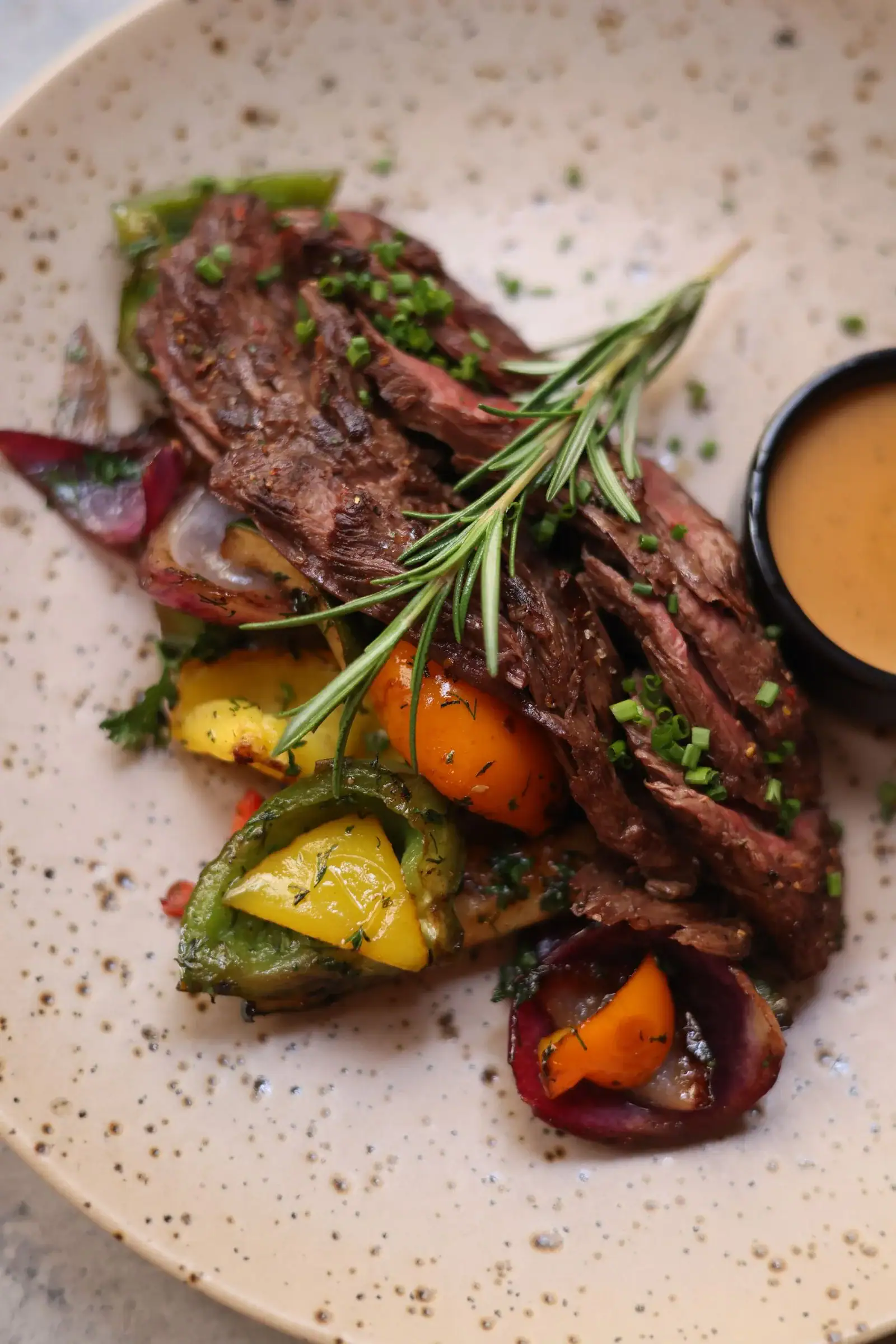 Sliced roasted meat with a rosemary sprig, roasted peppers and onions, and a small bowl of sauce on a speckled plate