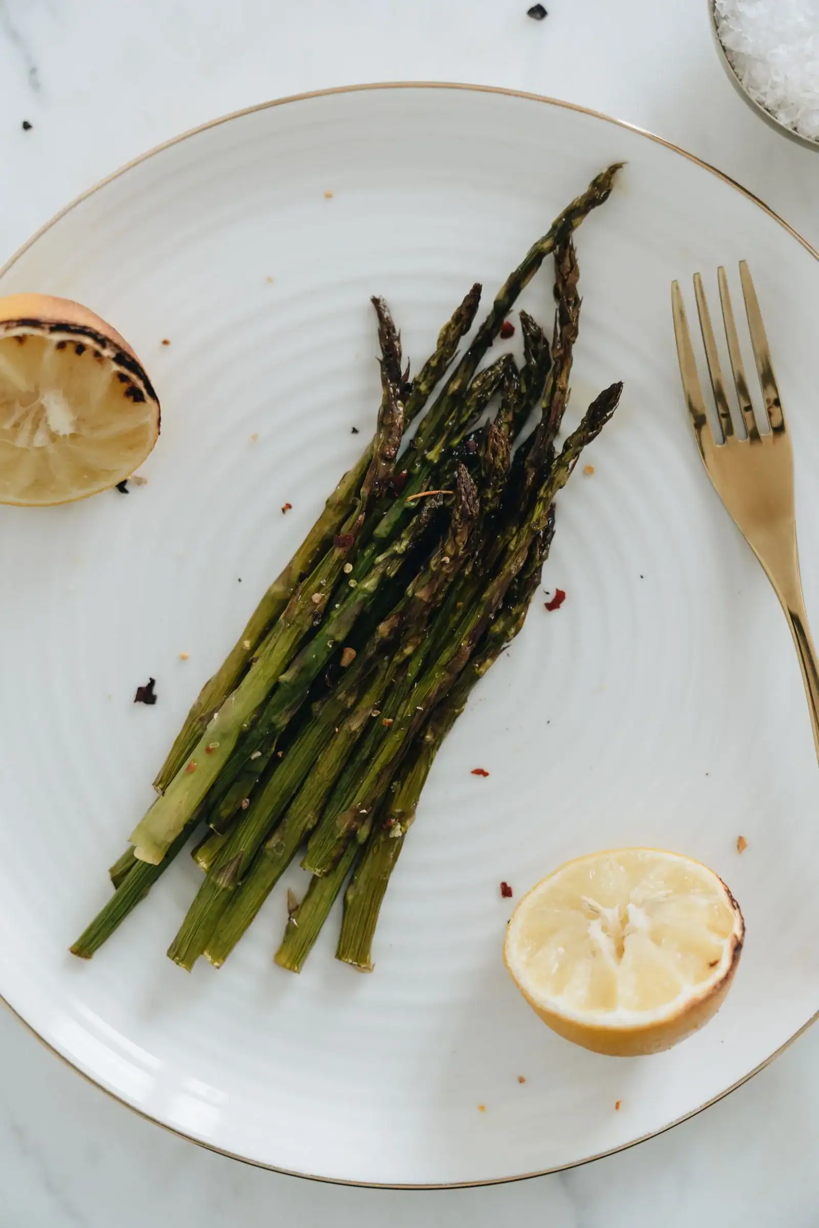 Roasted asparagus spears on a white plate beside a grilled lemon half and a fork