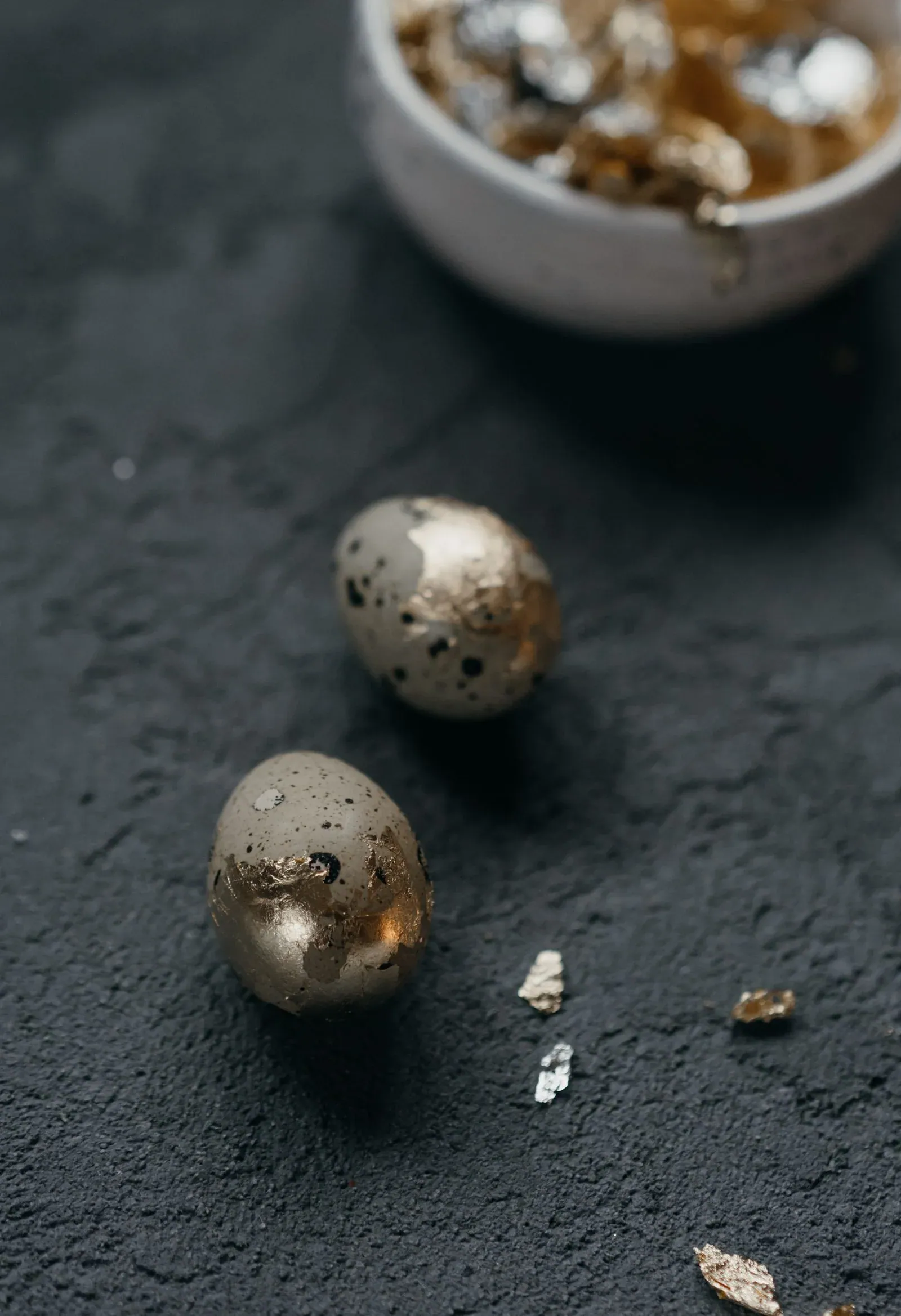 Two small eggs with gold leaf patches on a dark textured surface beside a bowl of gold flakes