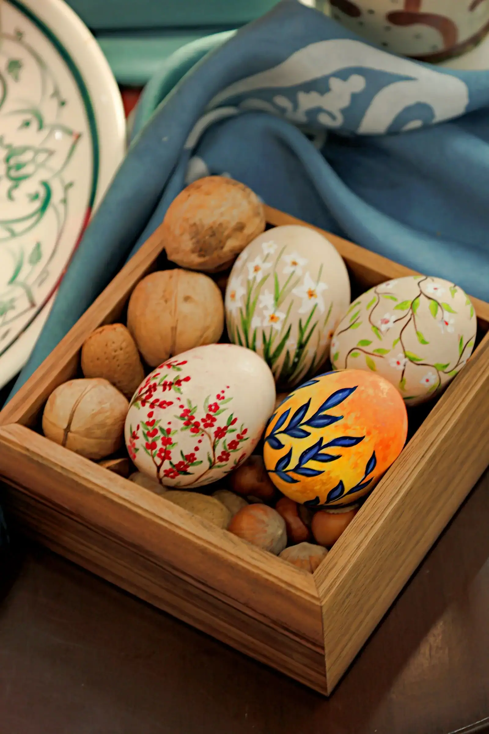 Hand-painted Easter eggs with red berry, white daisy and blue leaf designs in a wooden box with walnuts