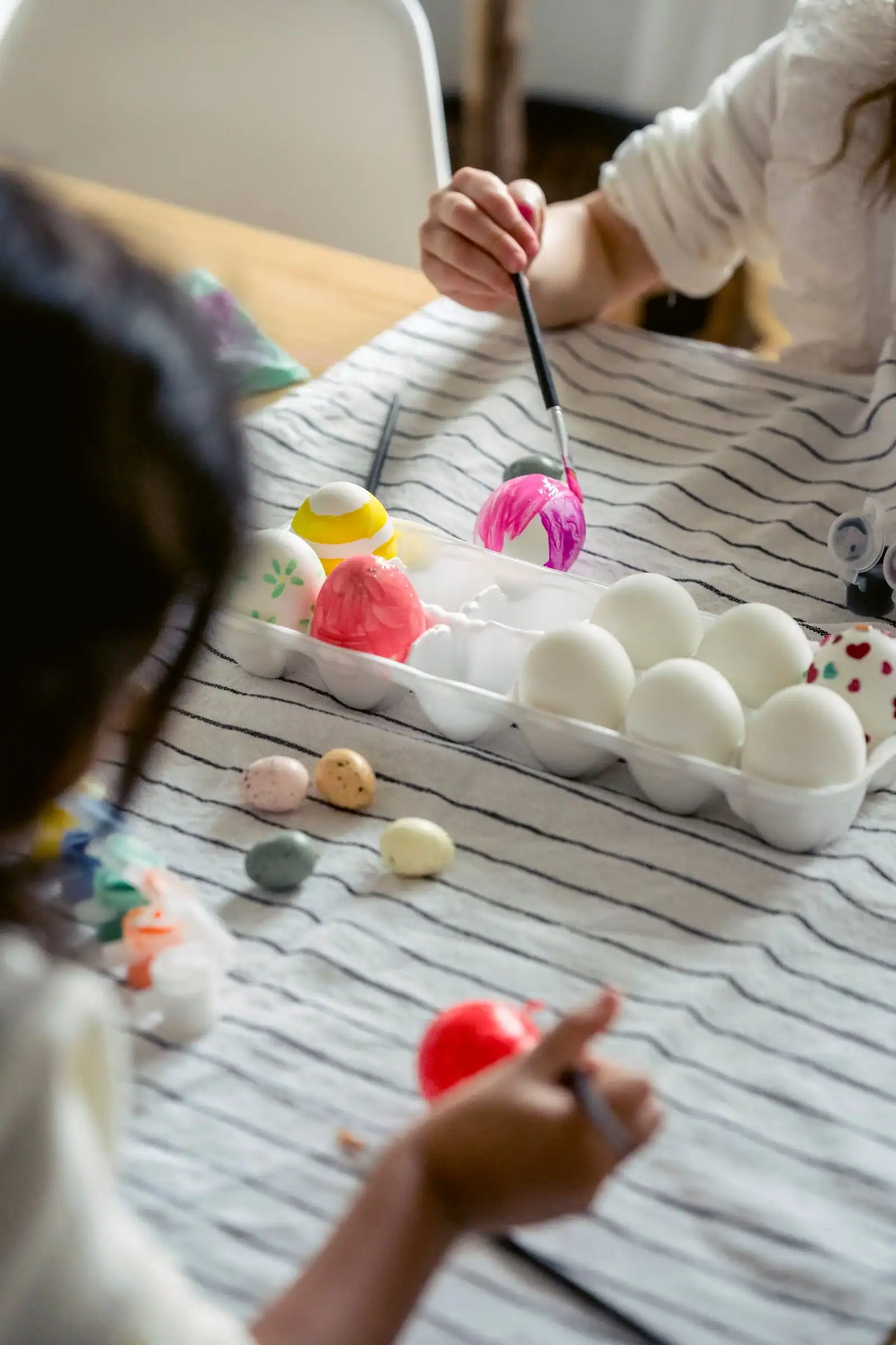 Children painting Easter eggs at a striped tablecloth with a tray of white eggs