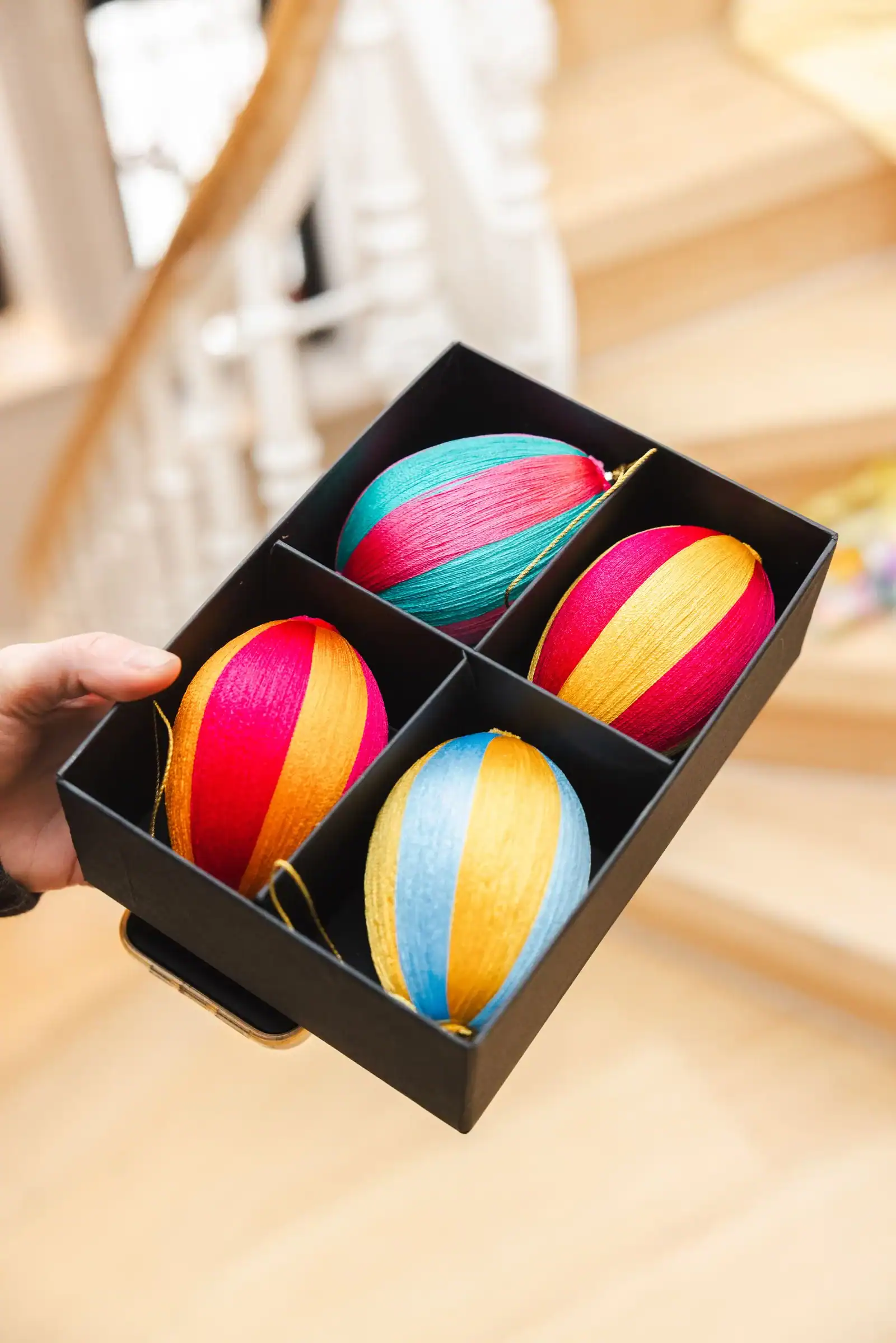 A hand holding a black box with four Easter eggs wrapped in red, orange, teal and blue thread