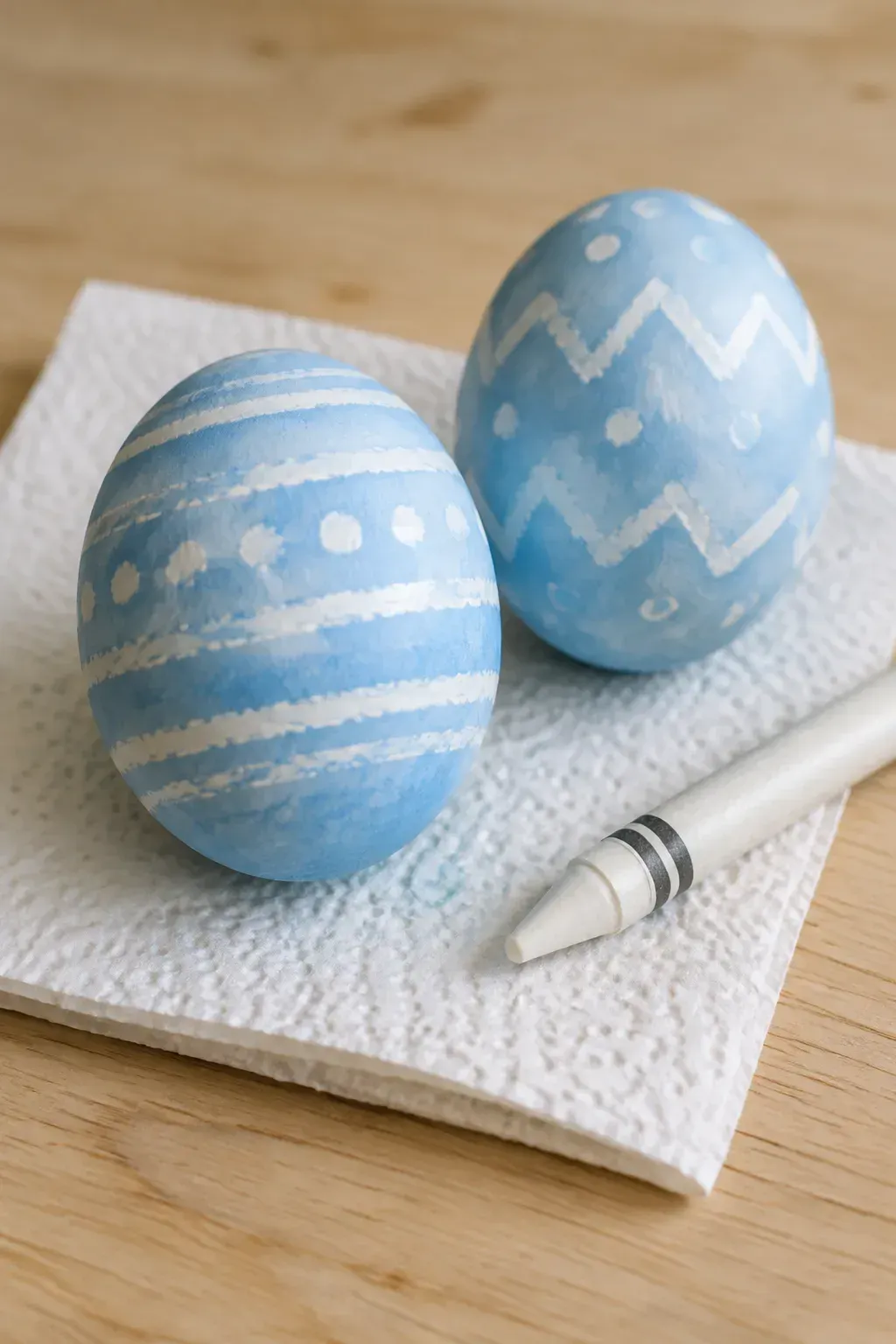 Two pale blue Easter eggs with white crayon stripes, zigzags and dots beside a white crayon on a paper towel