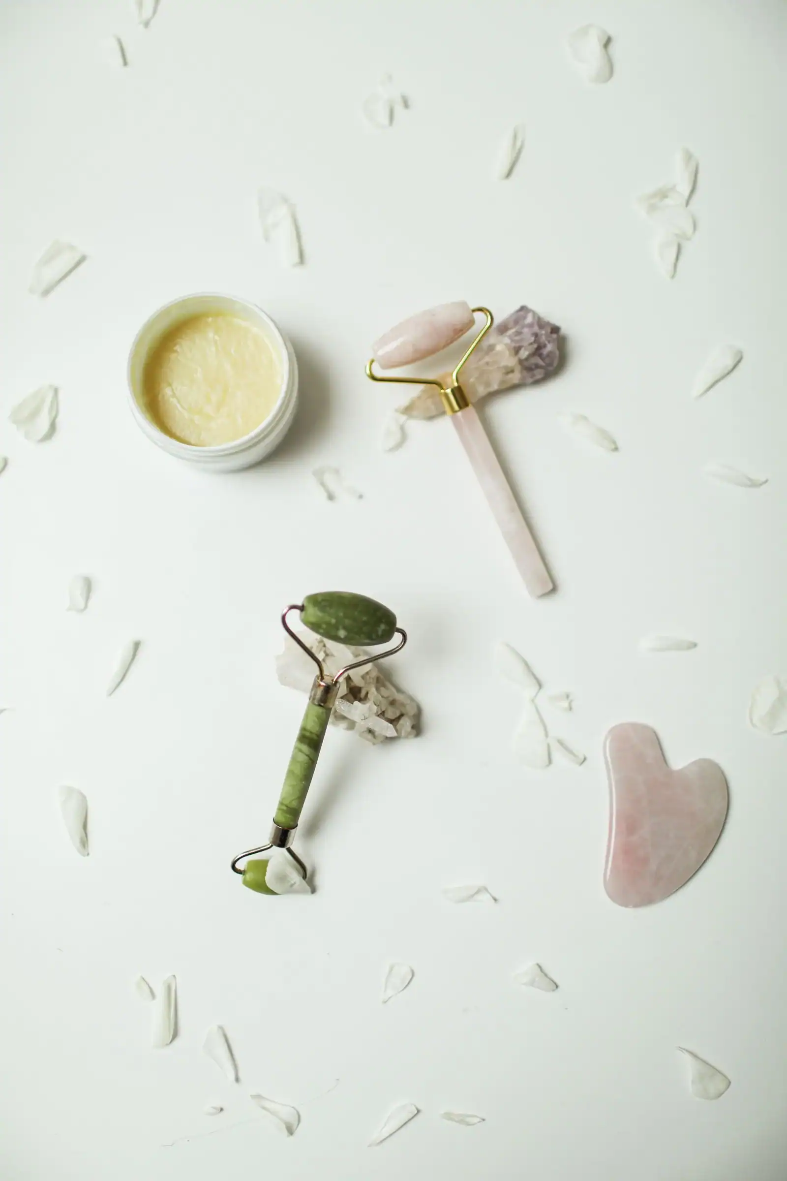 A flat lay of a jade roller, a rose quartz roller and a heart-shaped gua sha stone beside a jar of cream and petals