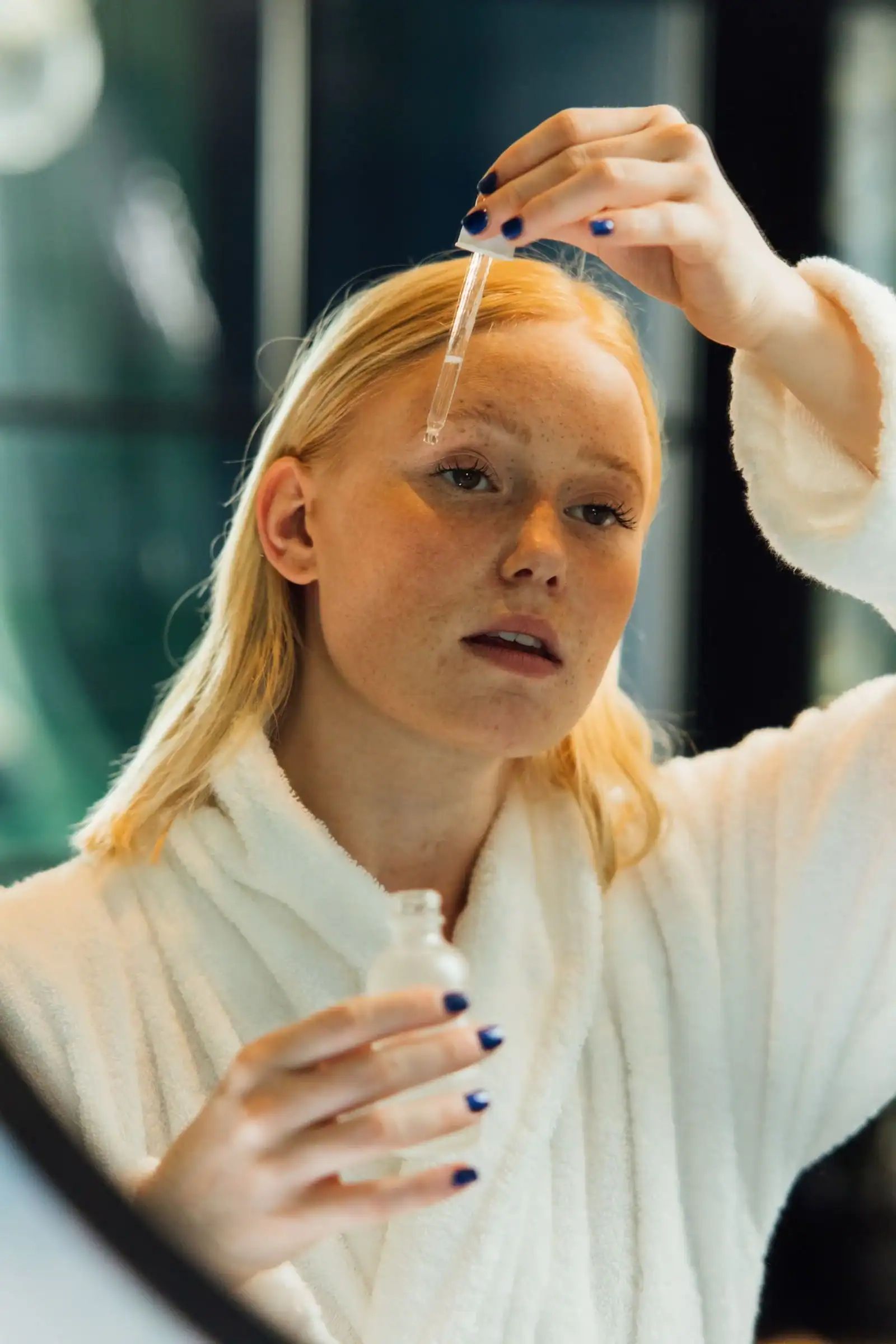 A woman in a white robe applying skincare serum to her forehead with a dropper in front of a mirror