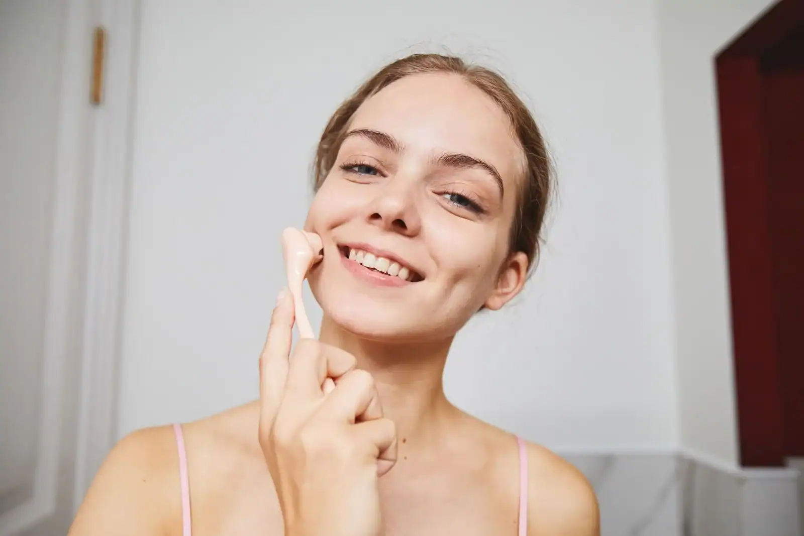 A smiling woman holding a pale pink facial massage tool against her cheek
