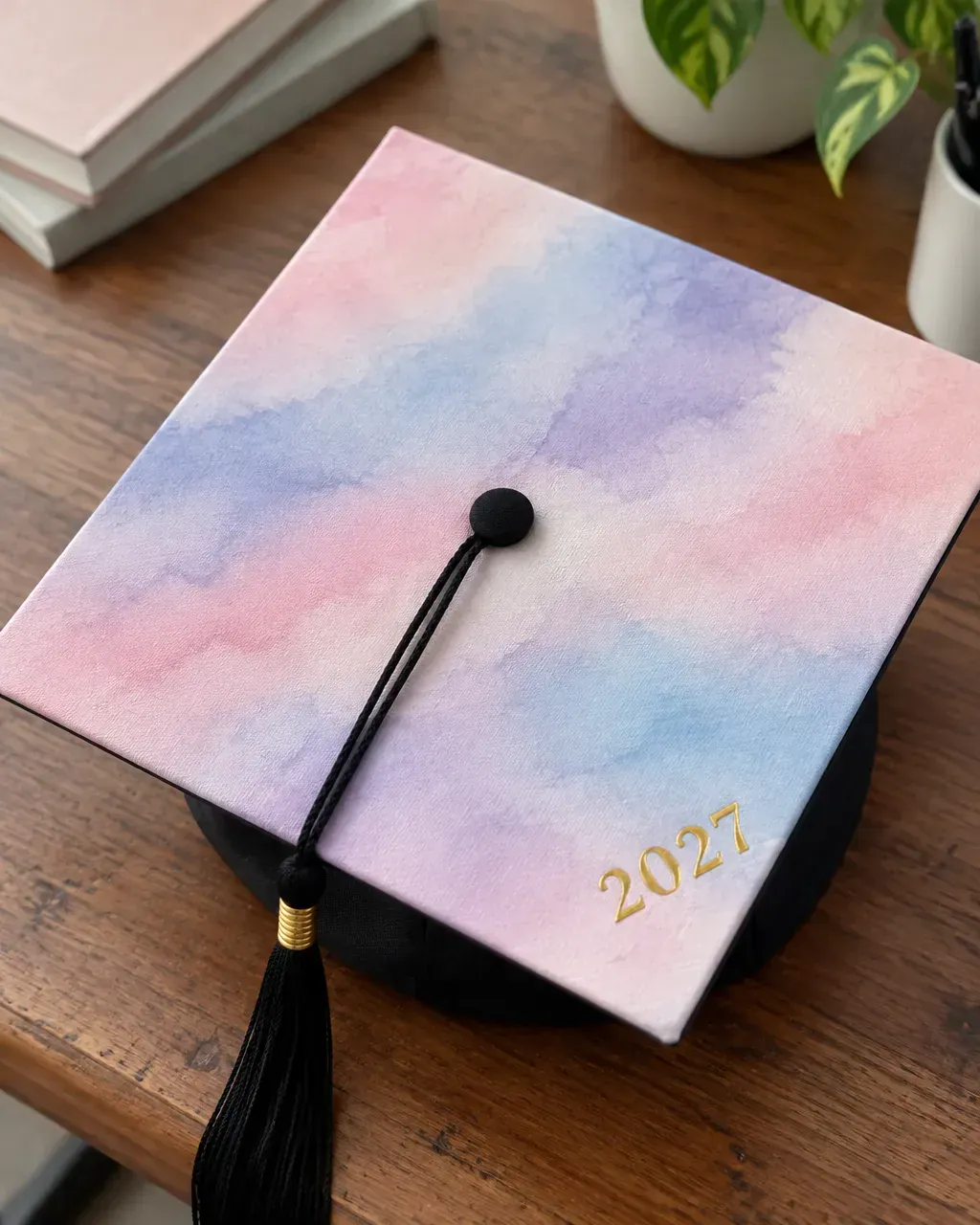 A graduation cap painted with a pastel watercolor wash in pink, lavender and blue with a small gold 2027