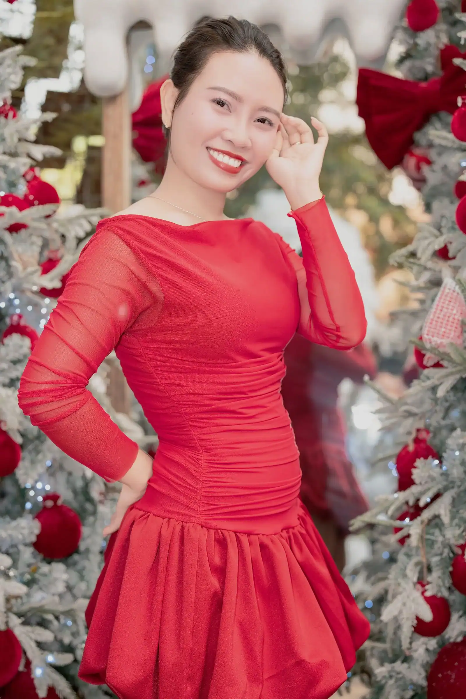 A woman in a red dress with sheer sleeves in front of a decorated Christmas tree