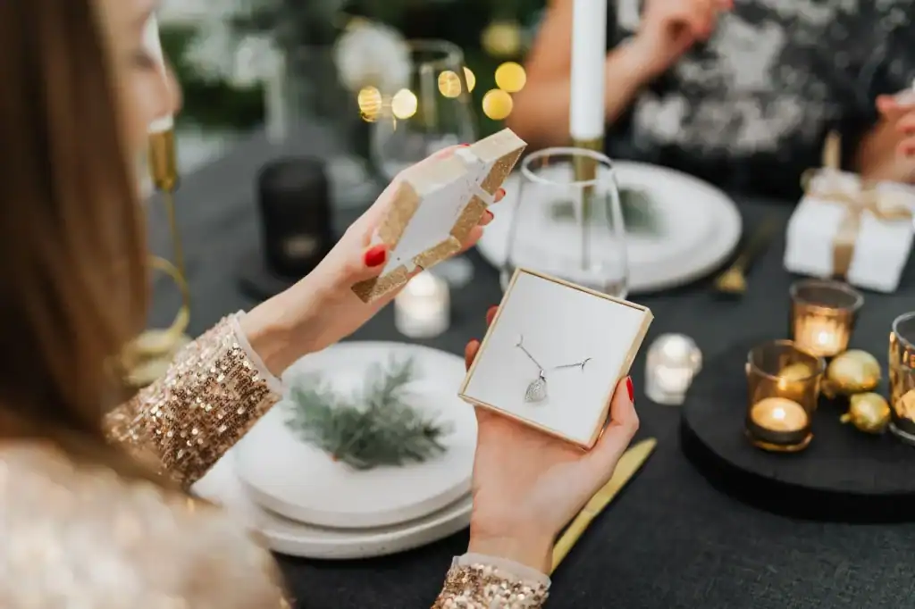 A woman opens a small box holding a pendant necklace at a candlelit holiday dinner table.