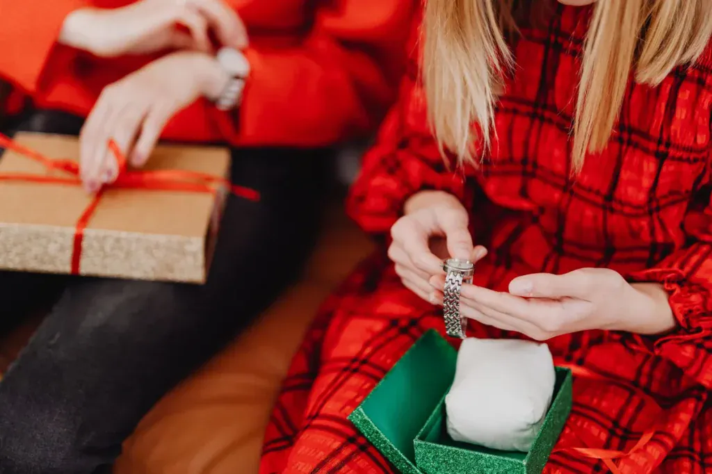 Hands in a red plaid dress lift a bracelet from a small green gift box next to a wrapped present.