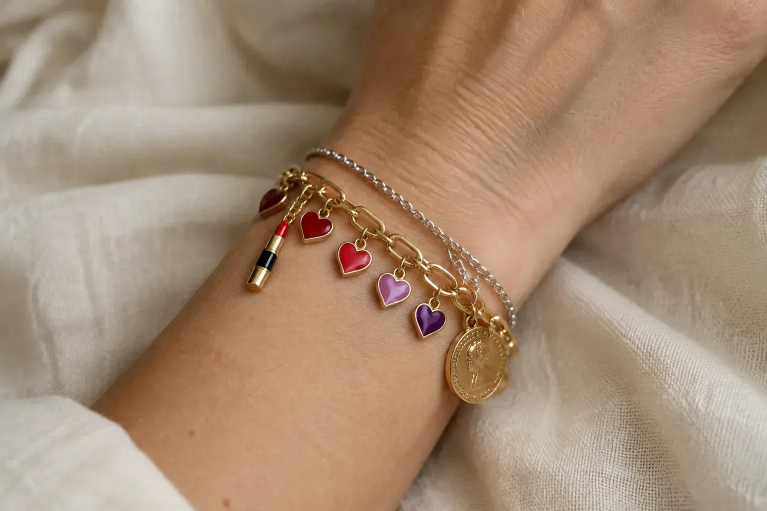 A wrist stacked with a gold charm bracelet carrying red and purple enamel heart charms, a lipstick charm and a coin charm, next to a thin silver chain.