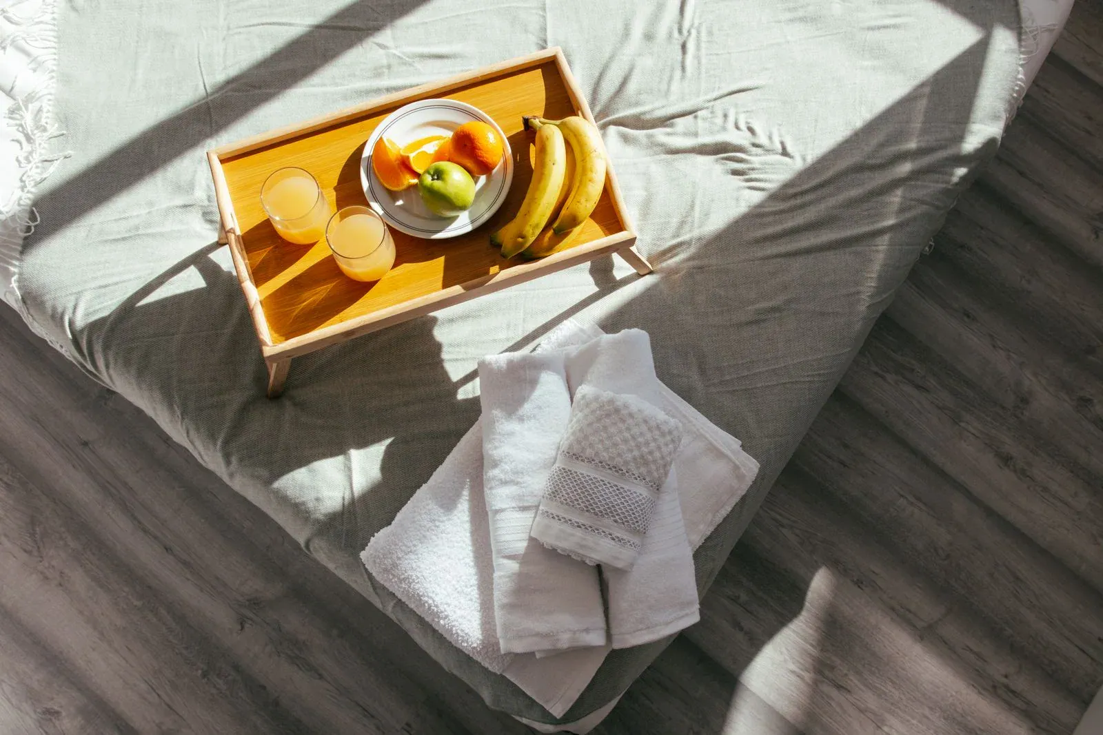 A wooden breakfast tray with fruit and juice on a bed, with folded white towels beside it