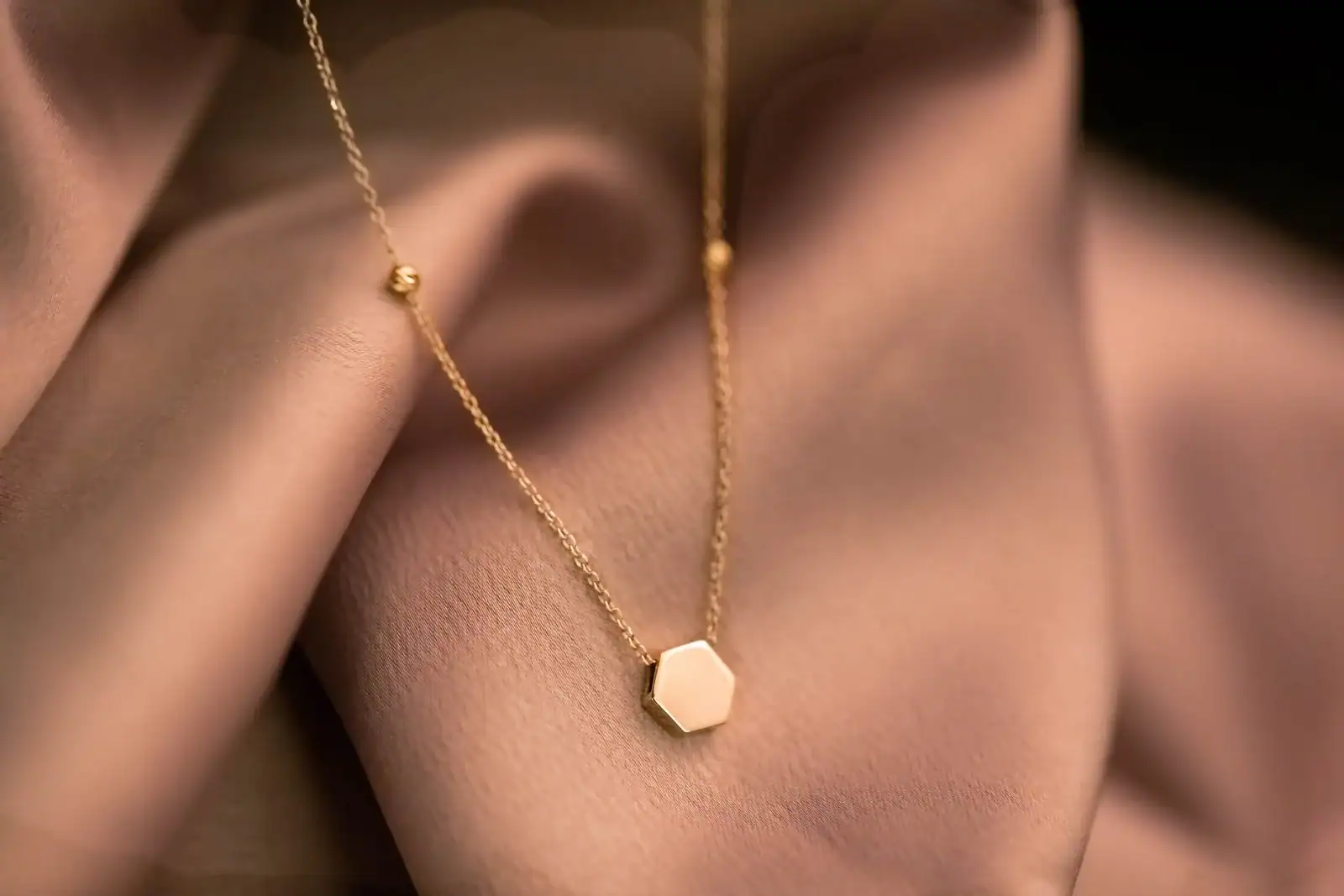 A thin gold necklace with a small hexagon pendant lying on dusty-pink satin