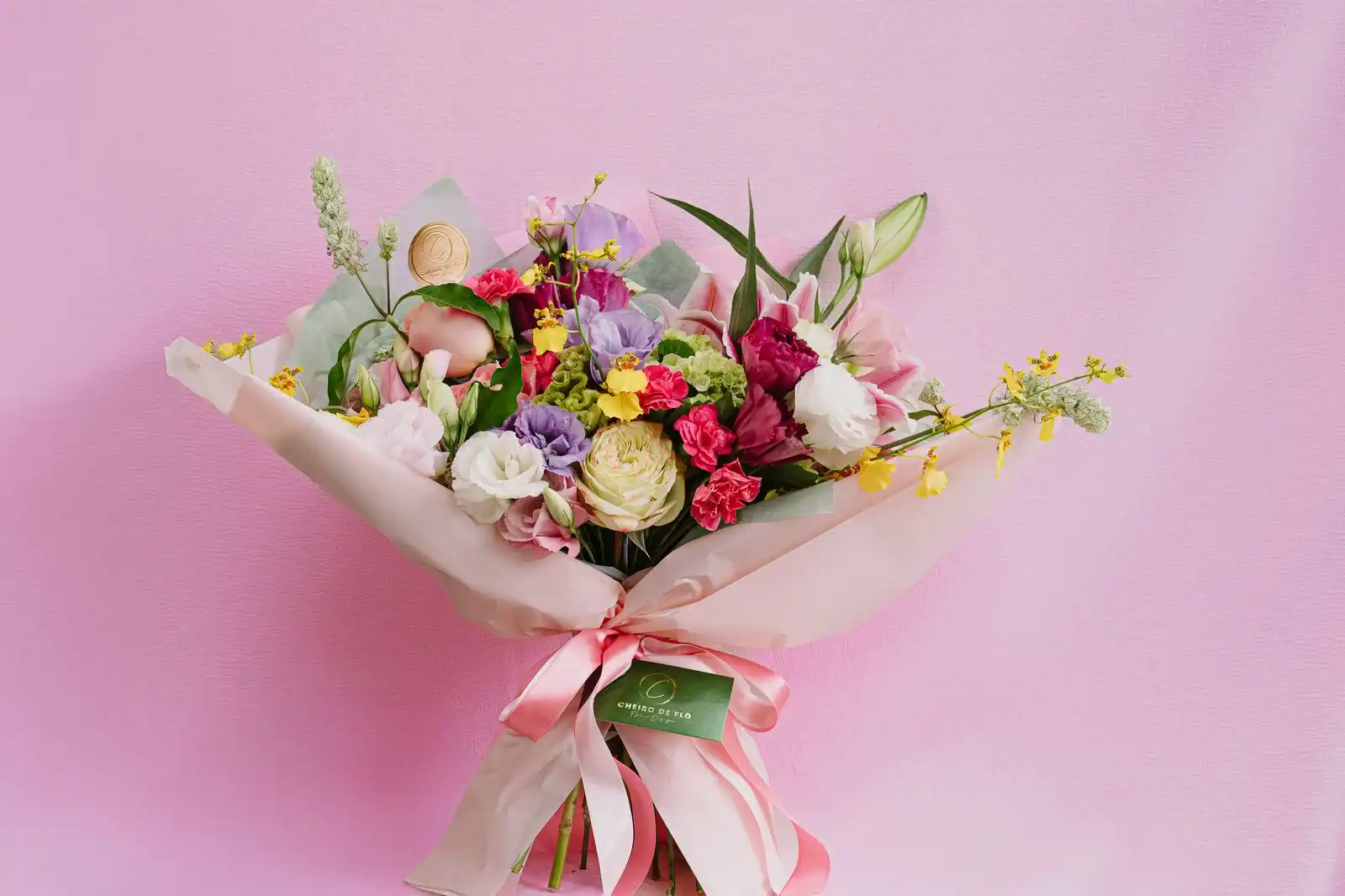 A wrapped bouquet of roses, lilies and spring flowers tied with a pink ribbon on a pink background