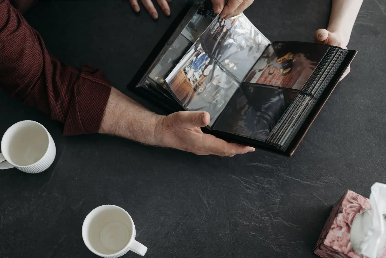 Hands holding an open photo album on a dark table beside two white coffee cups