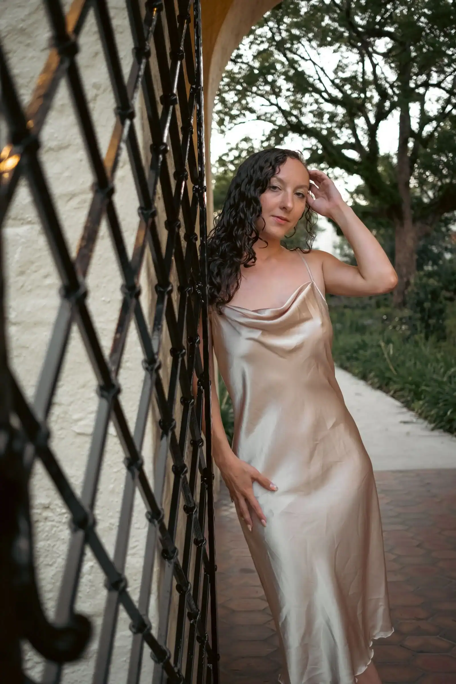 A woman in a long champagne satin slip dress leans on a lattice gate