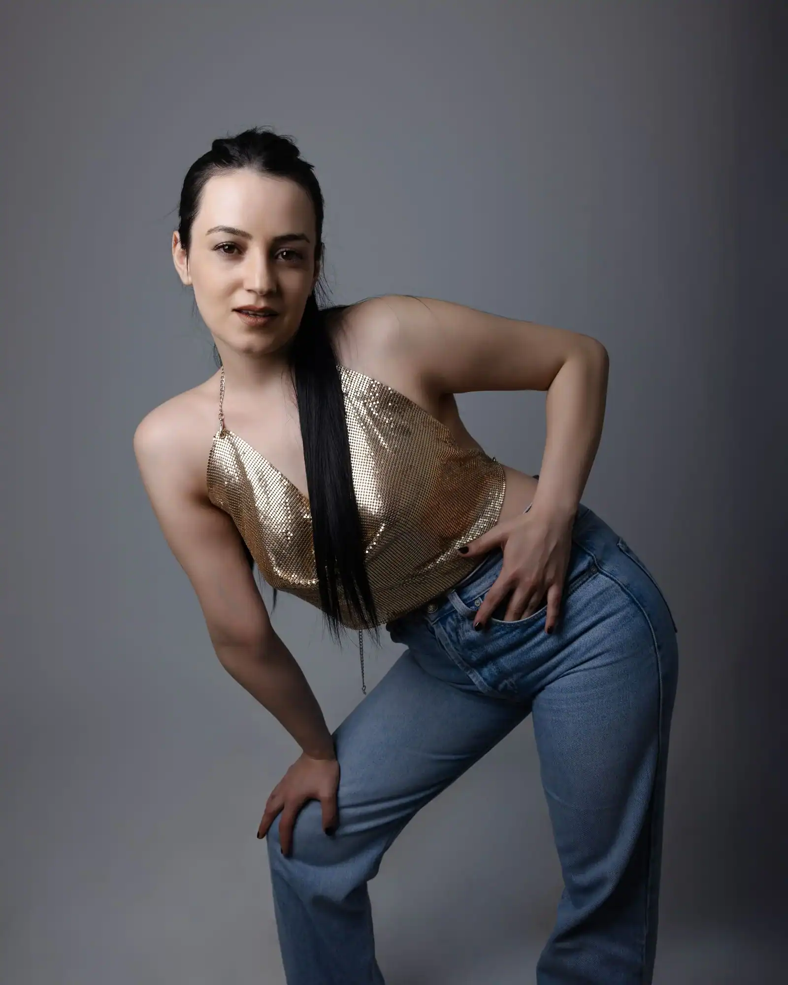 A woman in a gold satin top and light blue jeans poses indoors