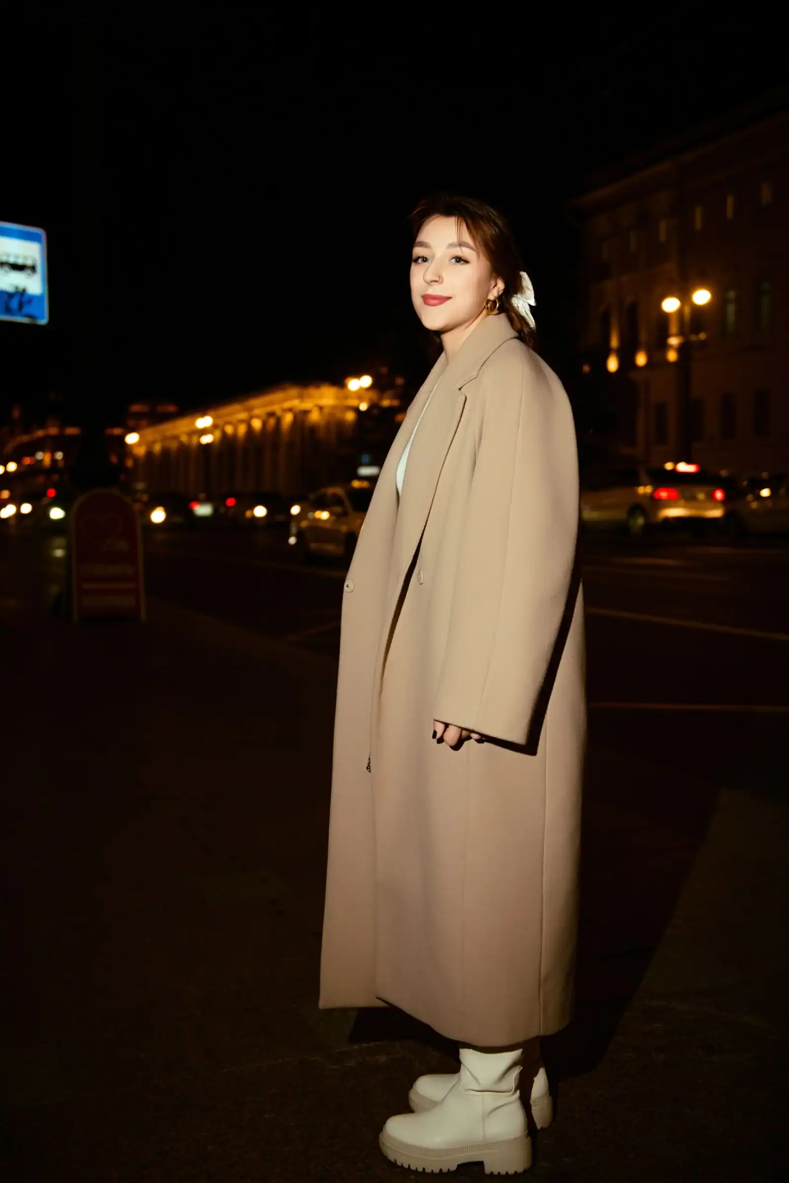 A woman in a long camel coat and white boots on a city street at night
