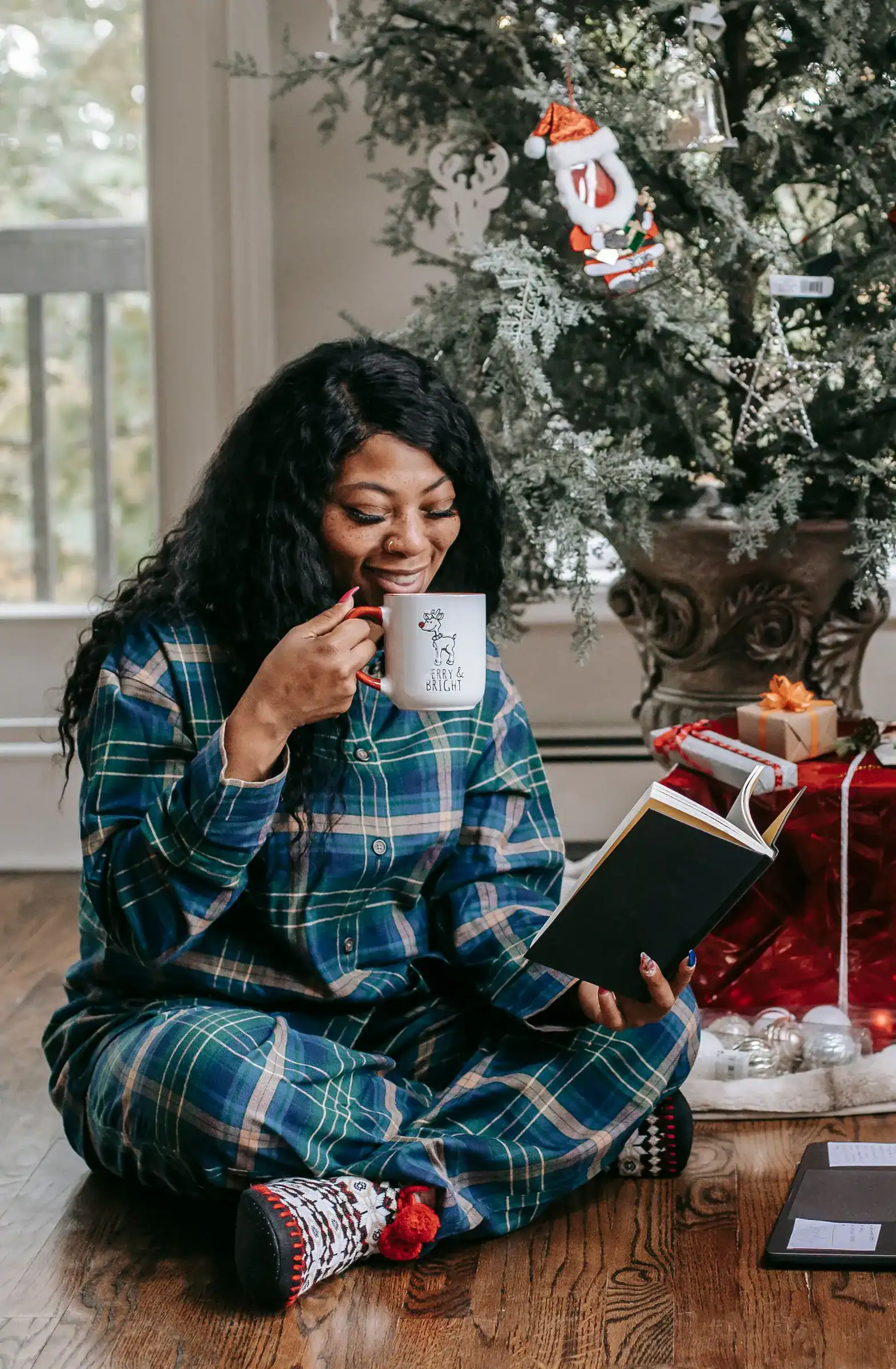 A woman in blue plaid pajamas sits on the floor with a mug and a book