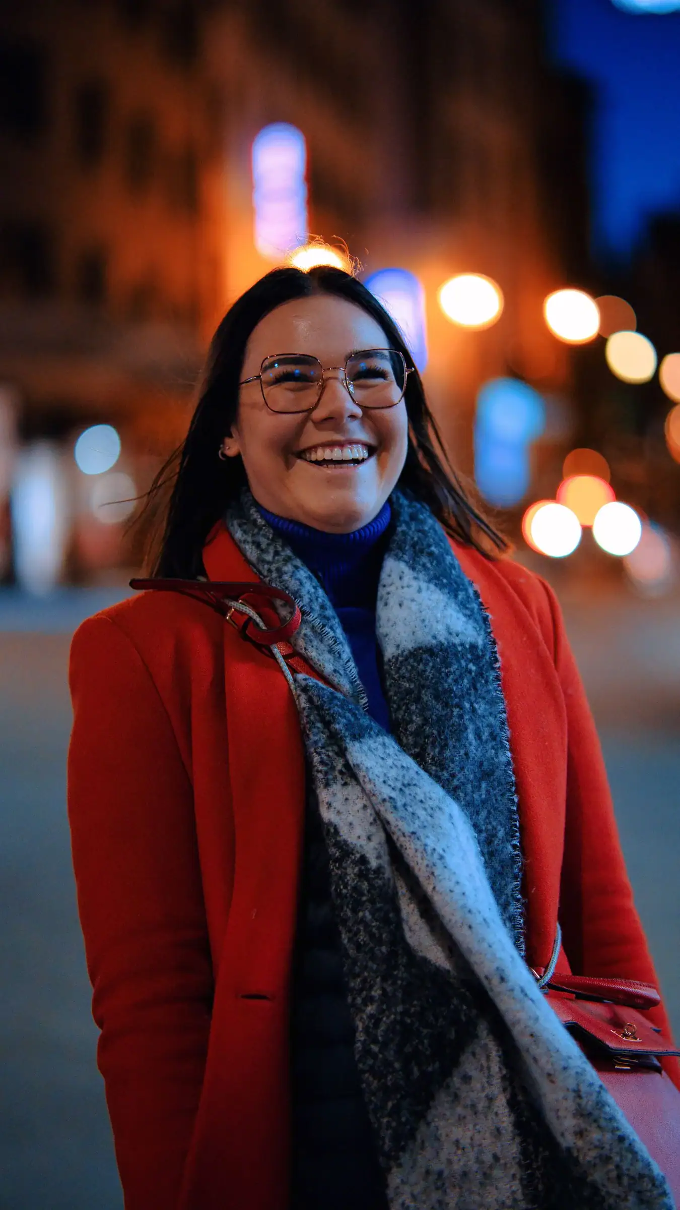 A smiling woman in a red wool coat and a large patterned scarf on a city street at night