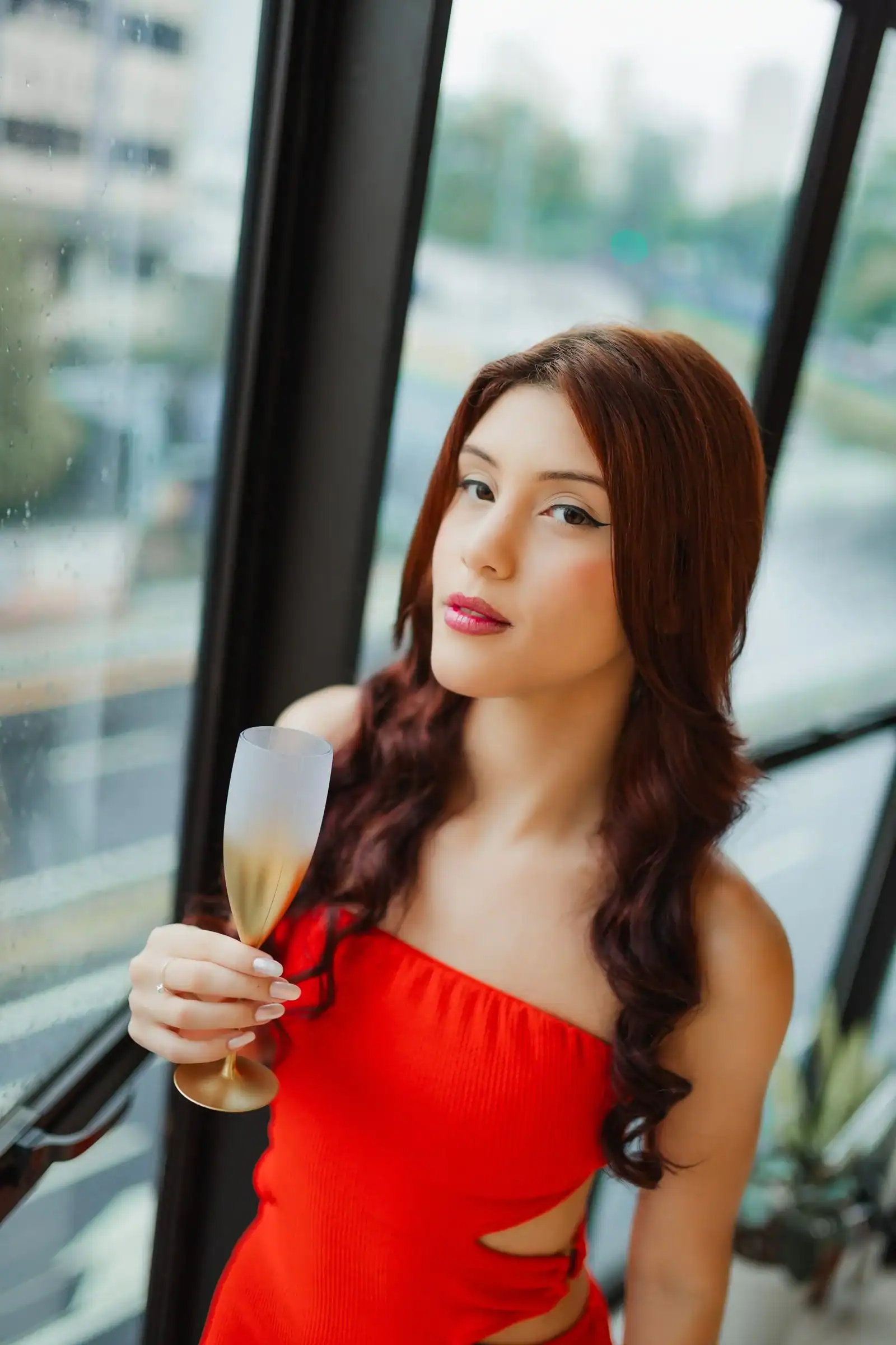A woman in a red one-shoulder dress holds a glass of champagne by a window
