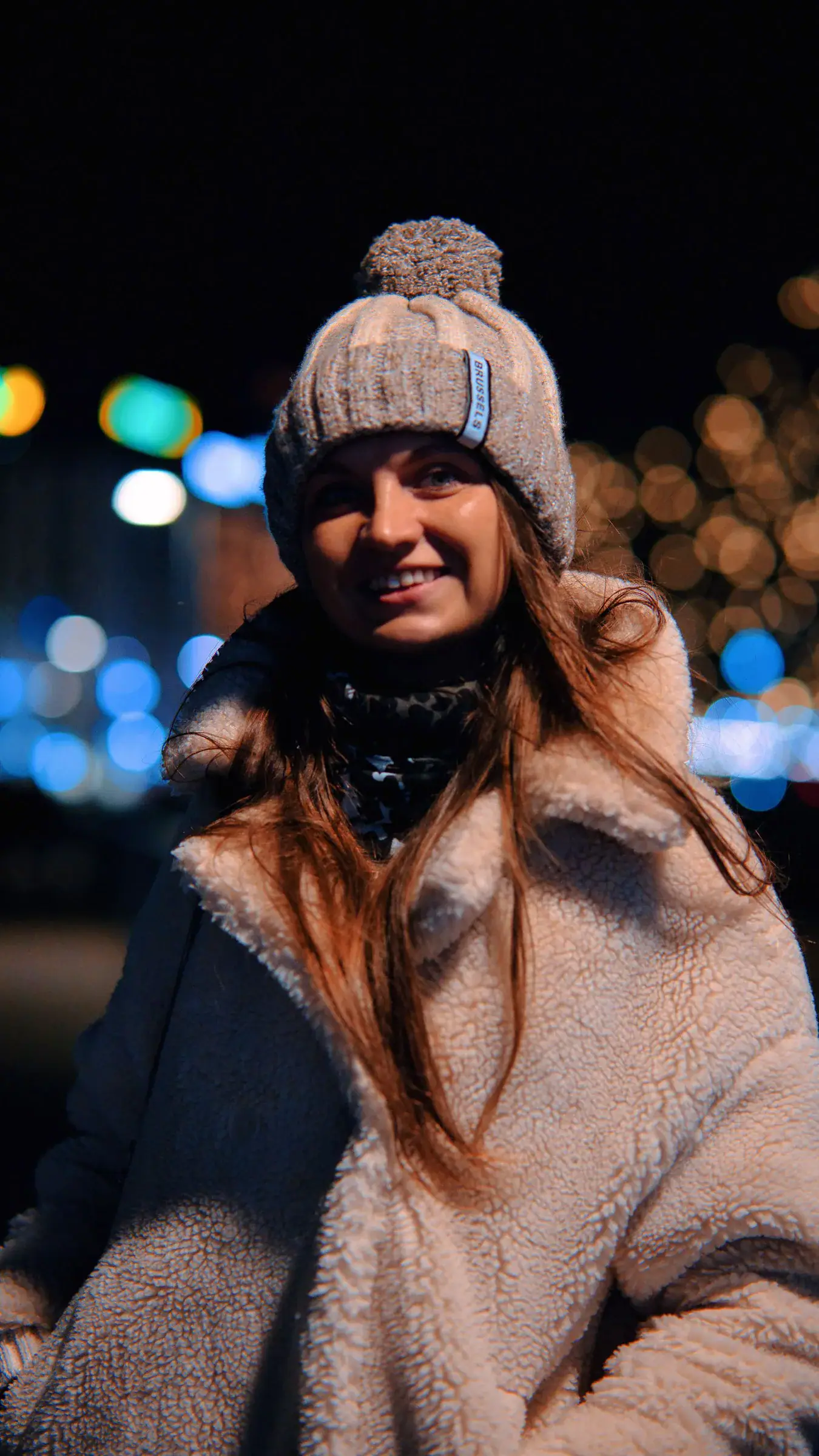 A smiling woman in a fluffy teddy coat and a knit beanie at night