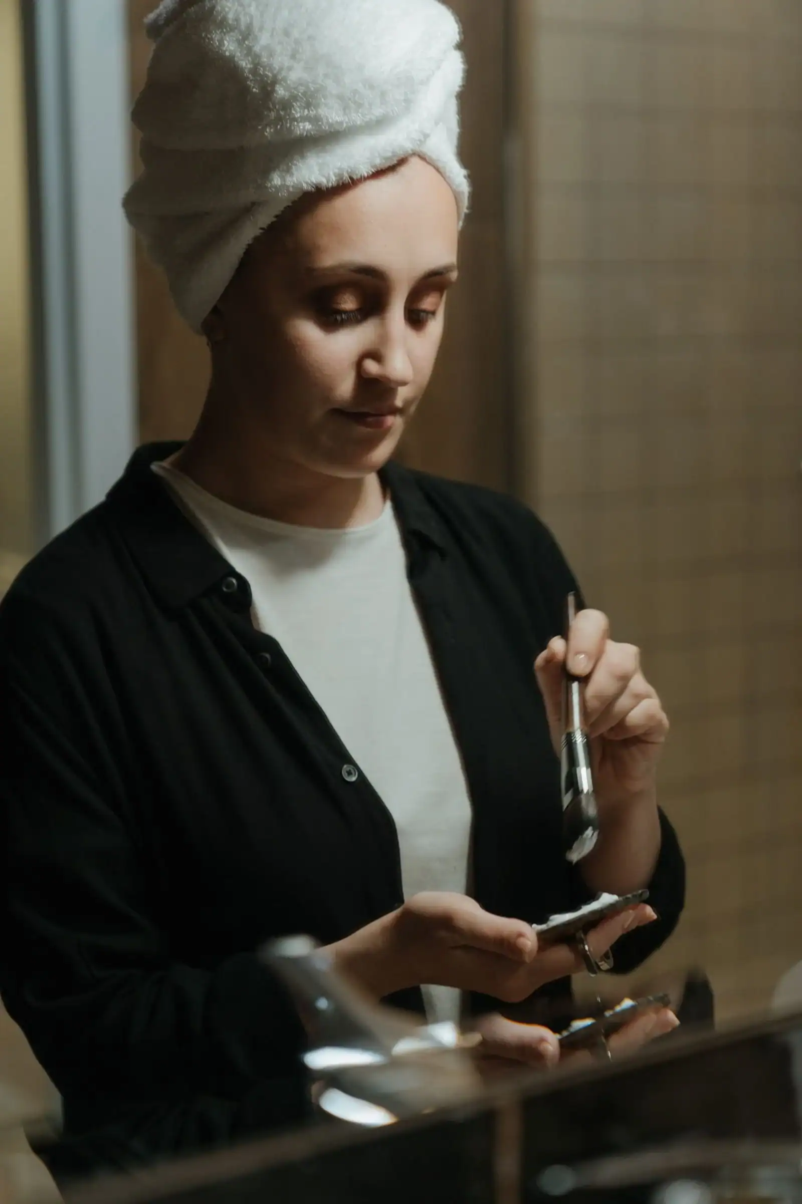 A woman with a towel on her head using a dropper at a bathroom mirror
