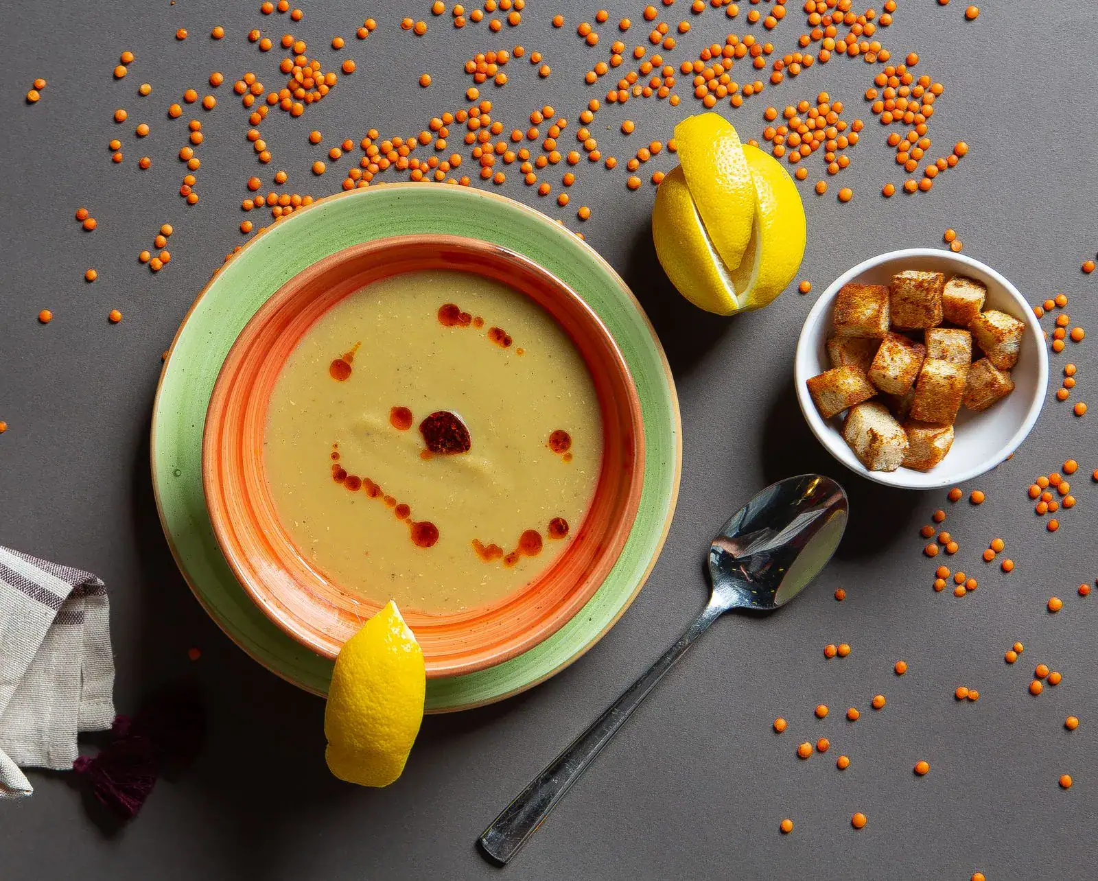 A bowl of red lentil soup with lemon wedges and croutons on a dark table
