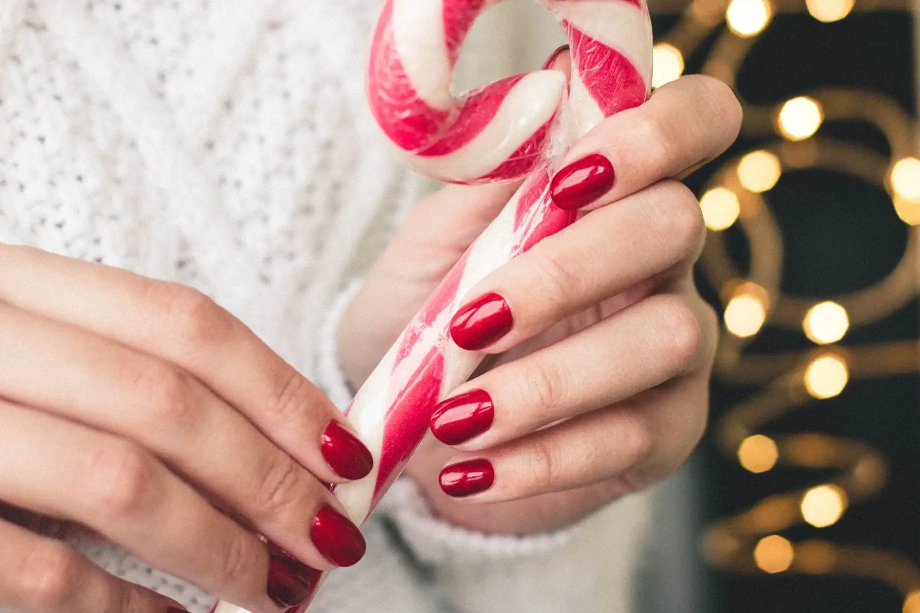 Red Christmas nails holding a candy cane in front of warm Christmas lights