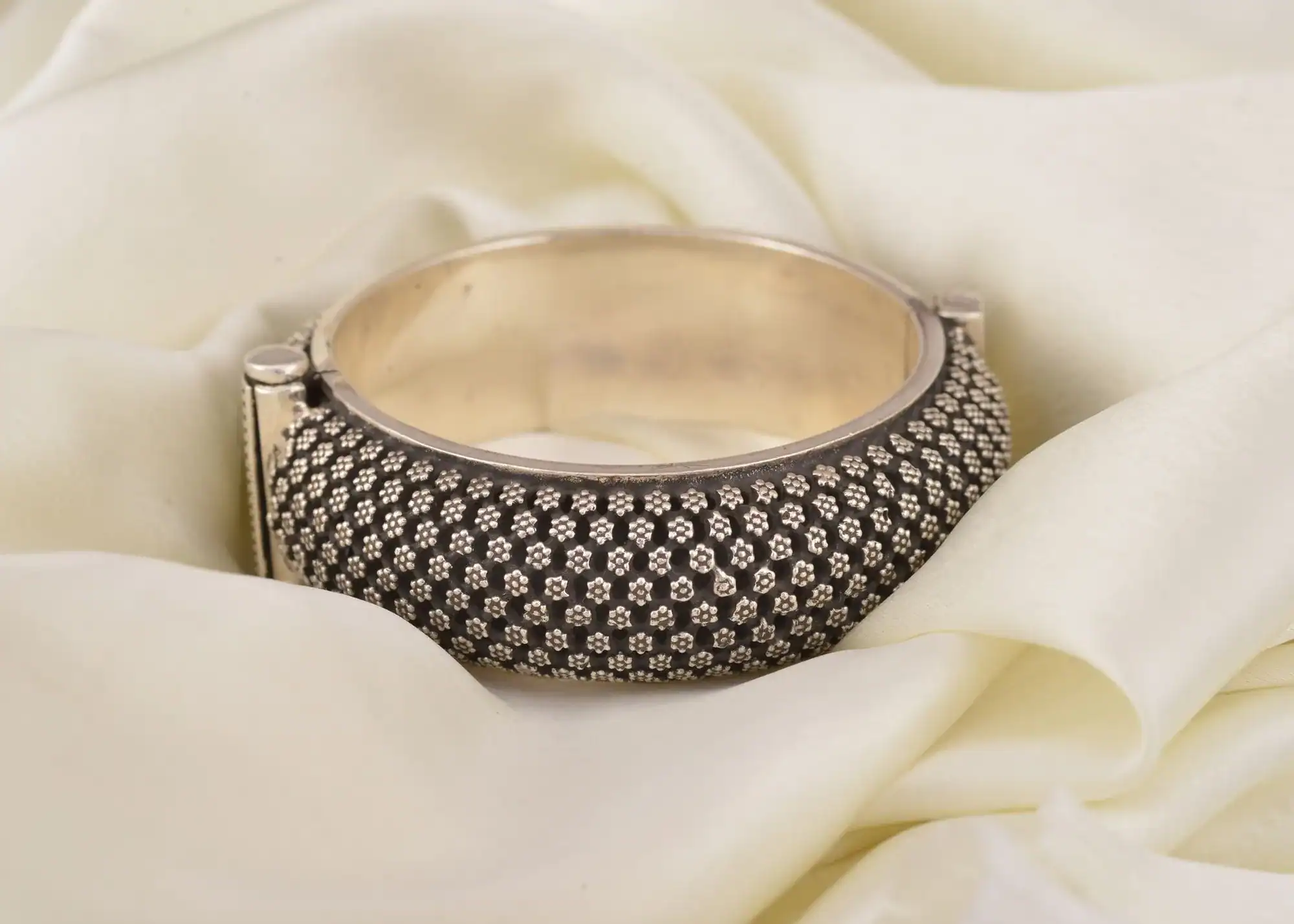 A wide sterling silver bangle with a blackened flower pattern resting on cream silk.