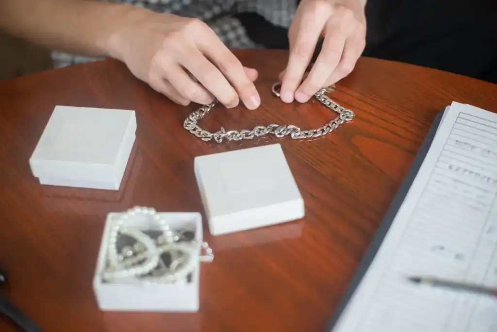 Hands laying out a silver chain necklace on a wooden table next to small white jewelry boxes.