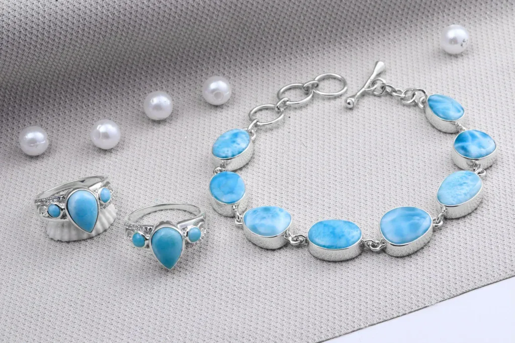 Silver rings and a bracelet set with pale blue larimar stones, with loose pearls on grey fabric.