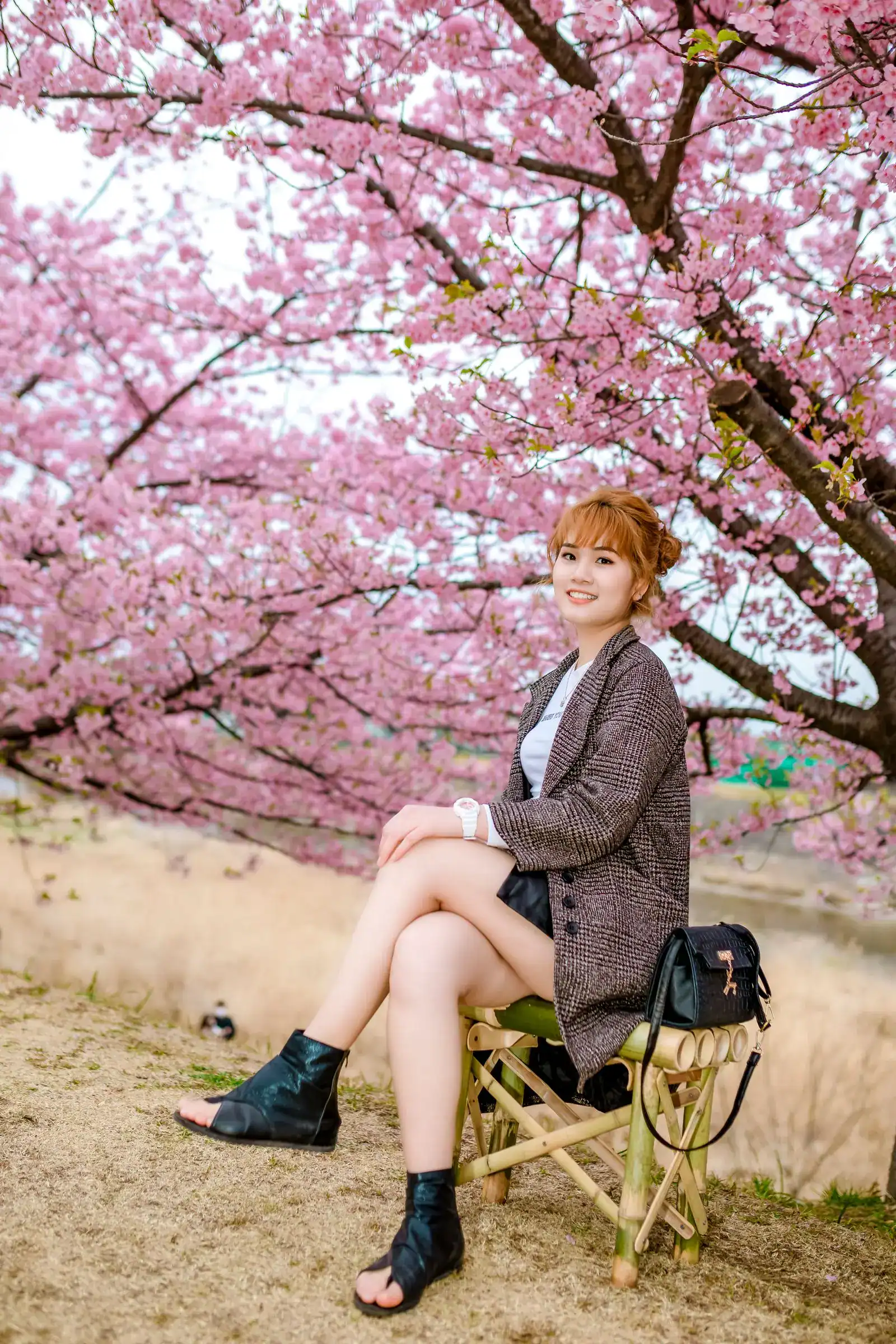 A woman in a grey blazer and boots sitting under cherry blossoms