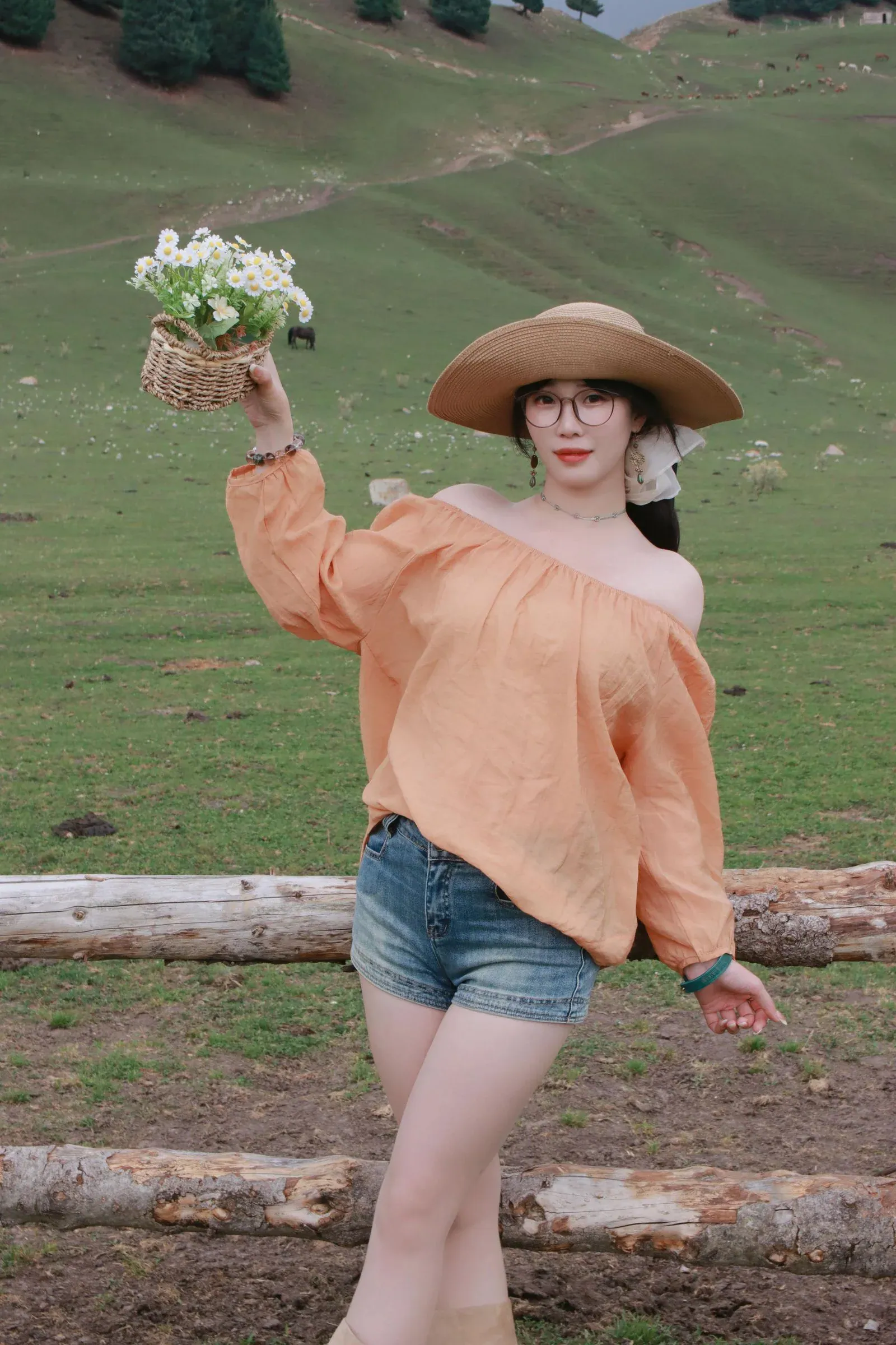 A woman in a peach off-shoulder top, denim shorts and a straw hat holding flowers