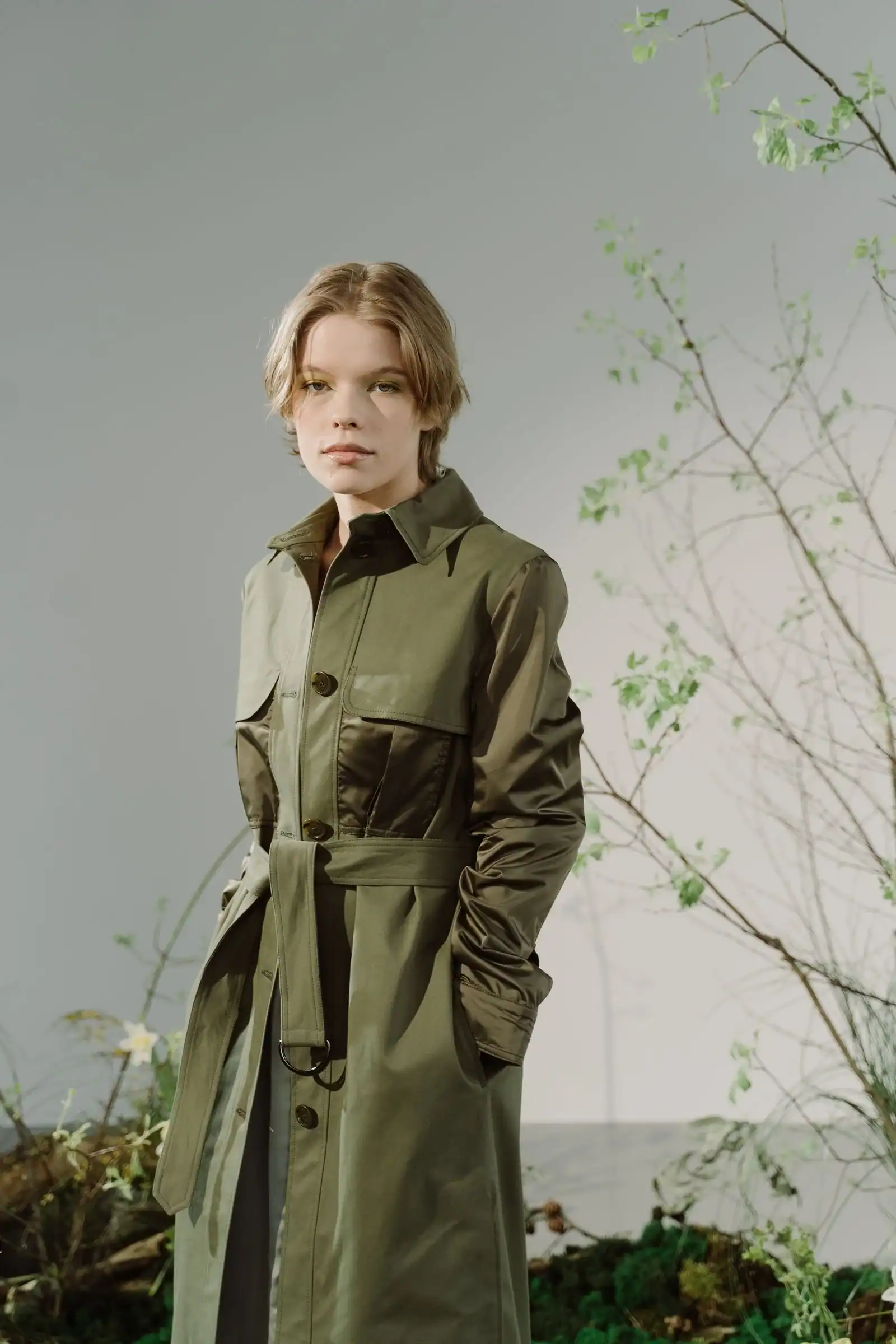 A woman in an olive green trench coat against a grey backdrop