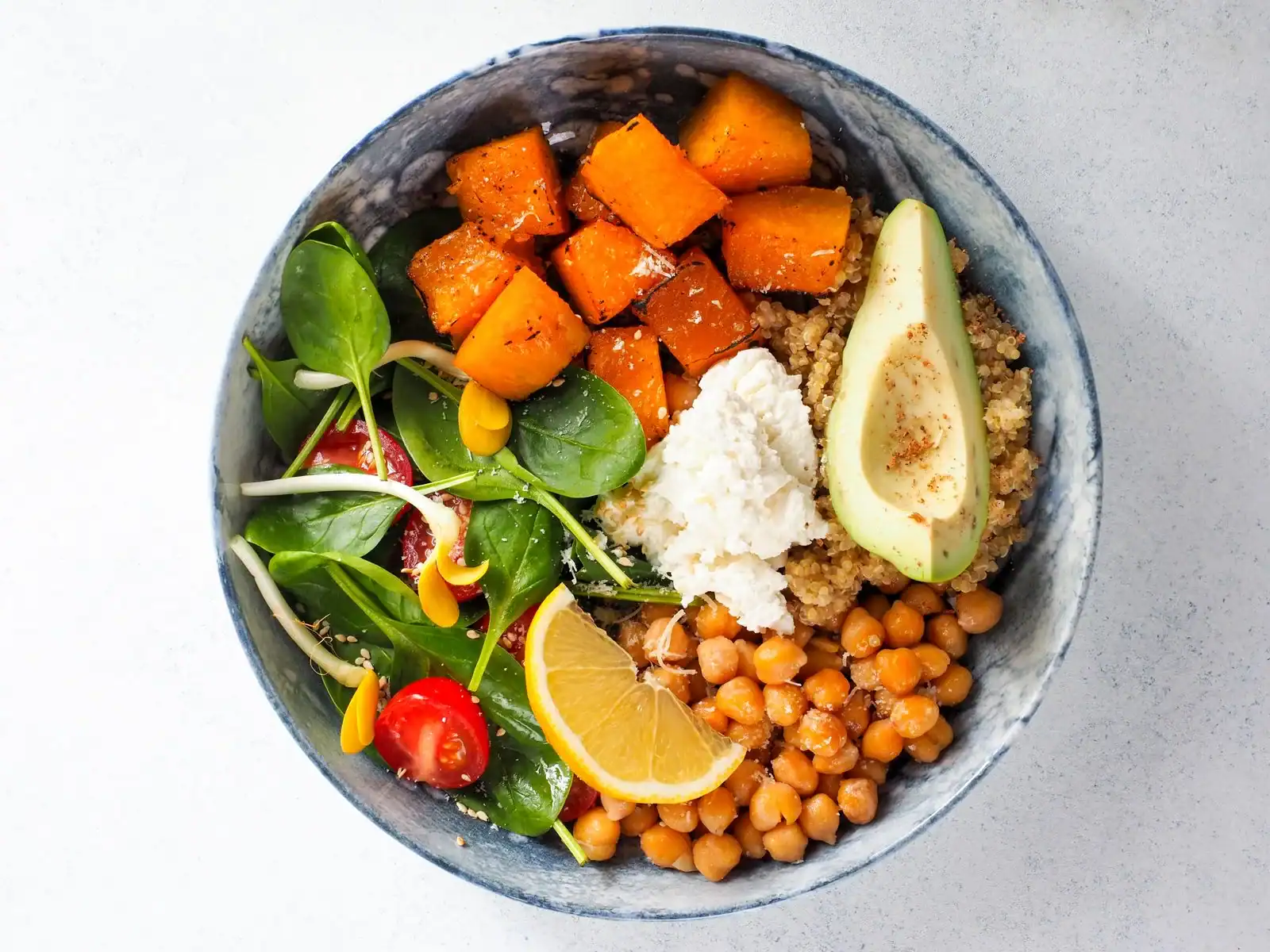 A bowl with roasted sweet potato, chickpeas, greens, avocado, a creamy topping and a lemon wedge